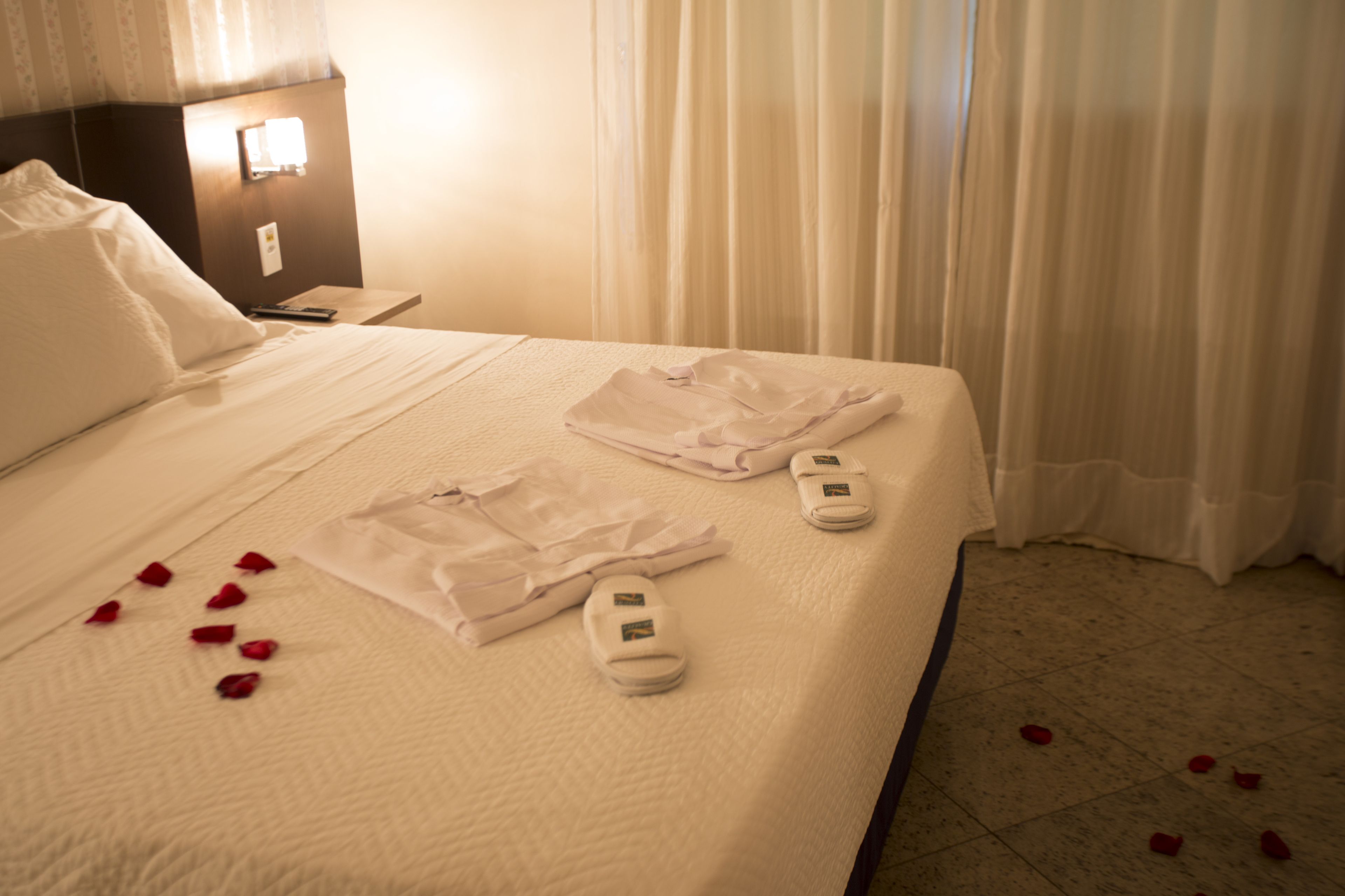 Una cama bien hecha con sábanas blancas, un par de zapatillas y pétalos de rosa esparcidos.