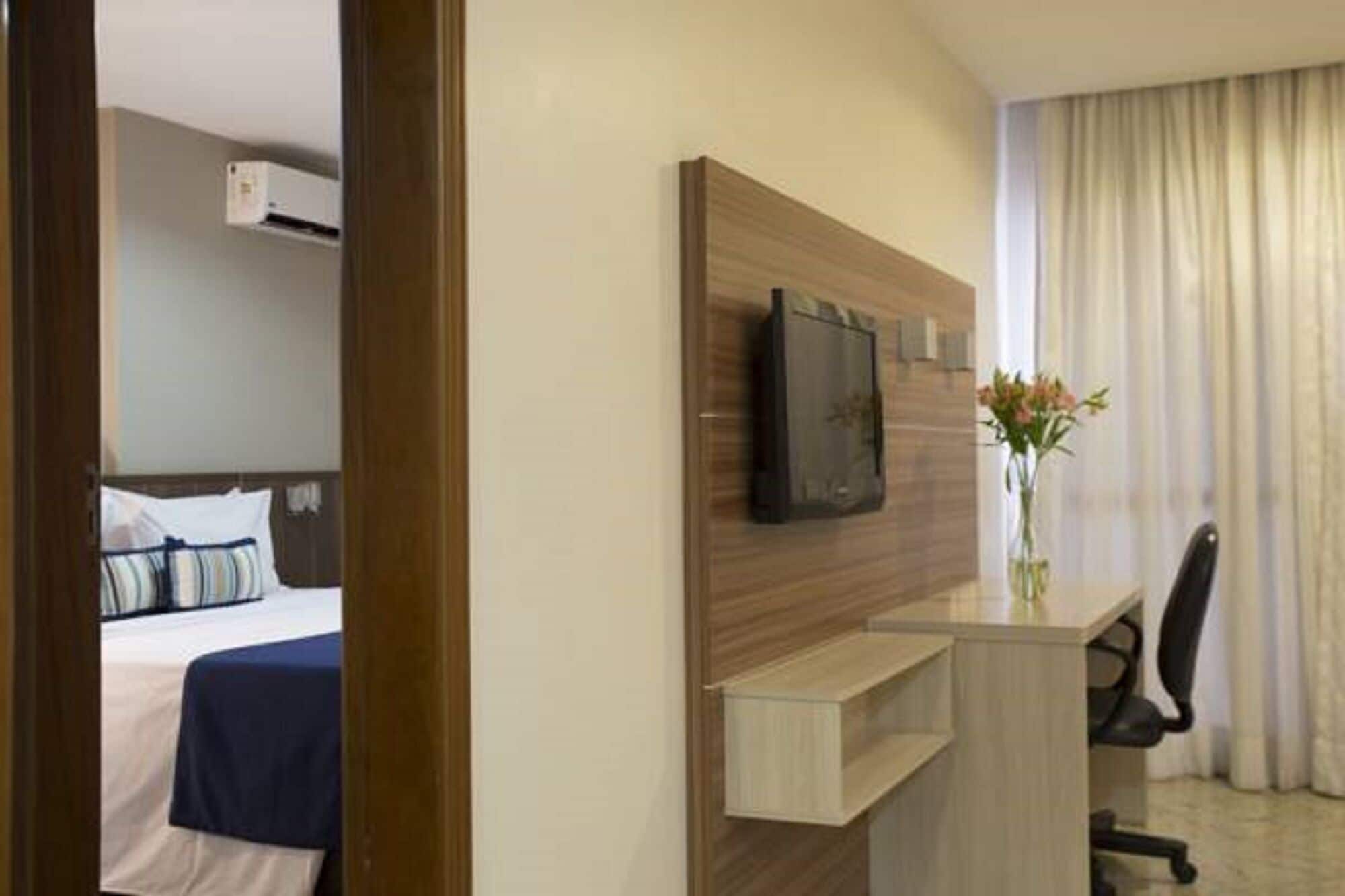 A hotel room with a bed, a desk with a chair, a flat-screen TV, and a vase with flowers.