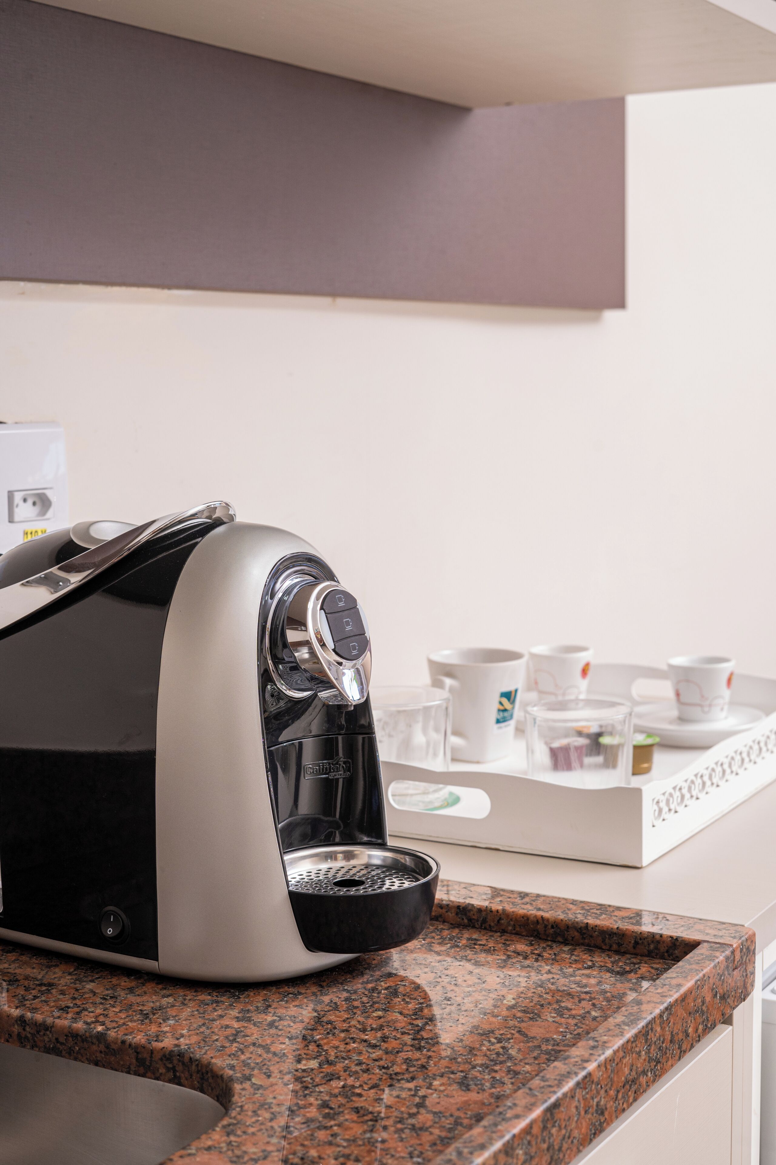 Uma cafeteira em uma bancada de granito, com uma bandeja de xícaras brancas e uma caixa de saquinhos de chá.