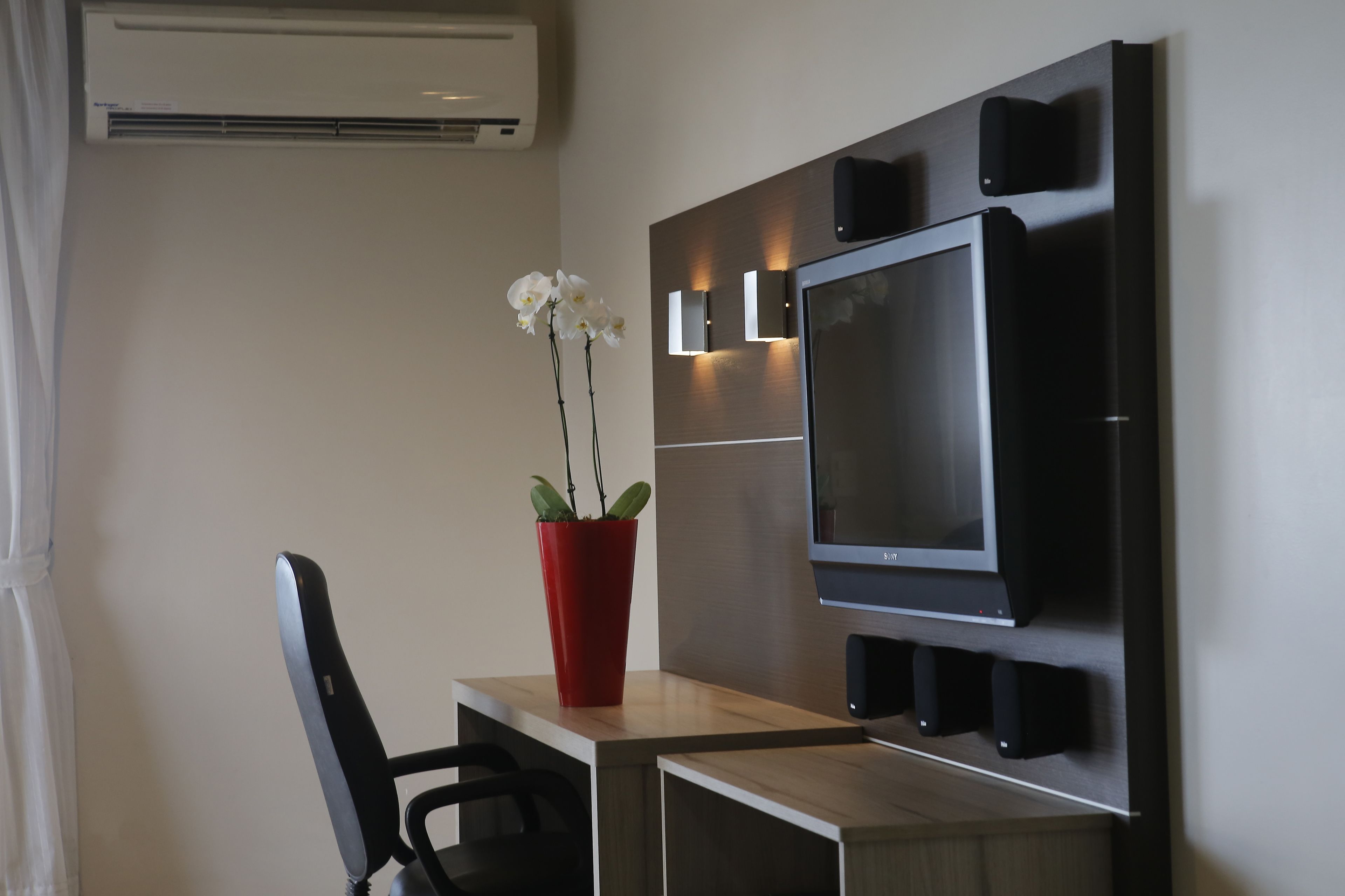 Una habitación de hotel moderna con un escritorio de madera, una silla negra, un florero rojo con flores blancas, un televisor de pantalla plana y altavoces integrados.