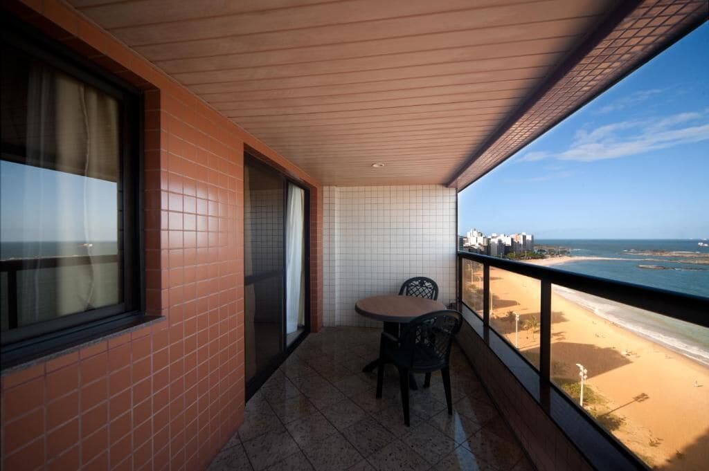 A balcony with a table and chairs, a view of the beach, and a cityscape in the background.