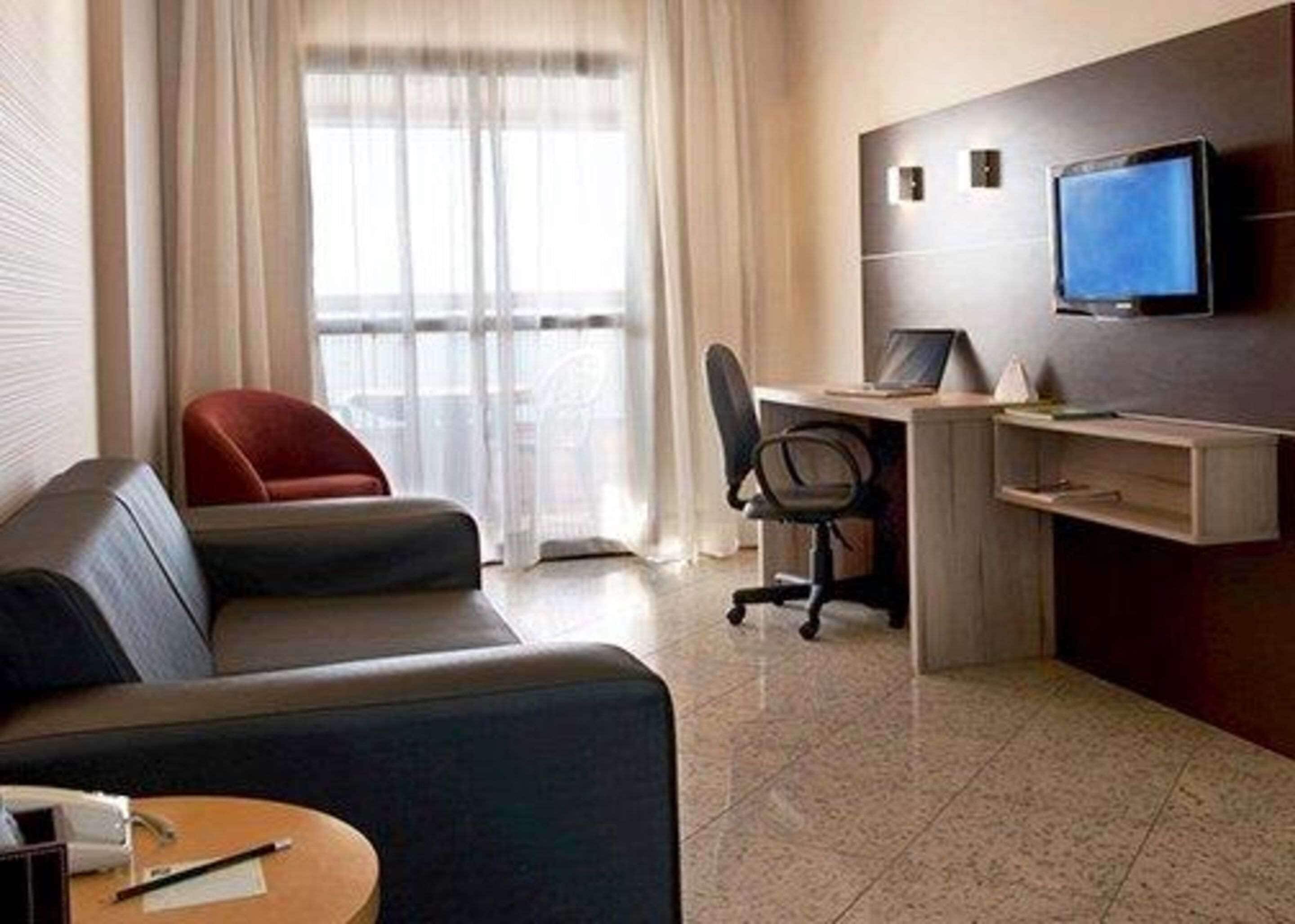 Una habitación de hotel moderna con sofá, escritorio con ordenador, televisor y ventana con cortinas.