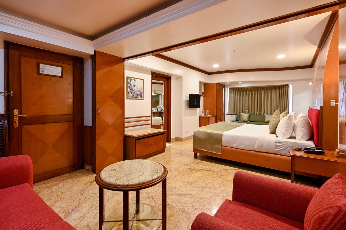 Quarto de hotel com uma cama, um espaço de estar com um sofá vermelho, uma mesinha e uma porta de madeira.