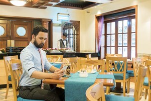 A man sitting at a table with a smartphone, a drink, and a plate of food in a restaurant with wooden furniture and large windows.