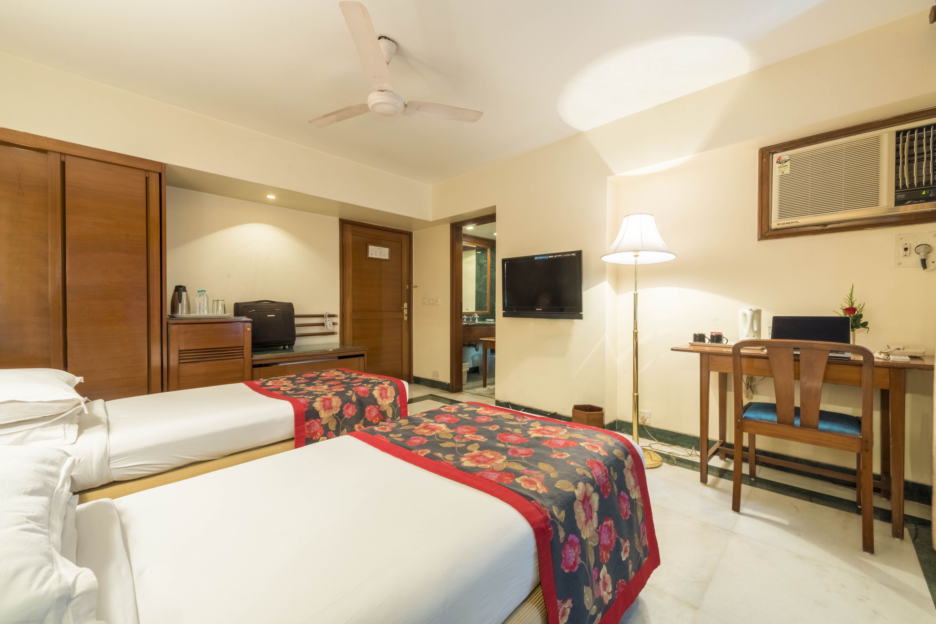 A hotel room with two beds, a desk, a chair, a television, and a ceiling fan.