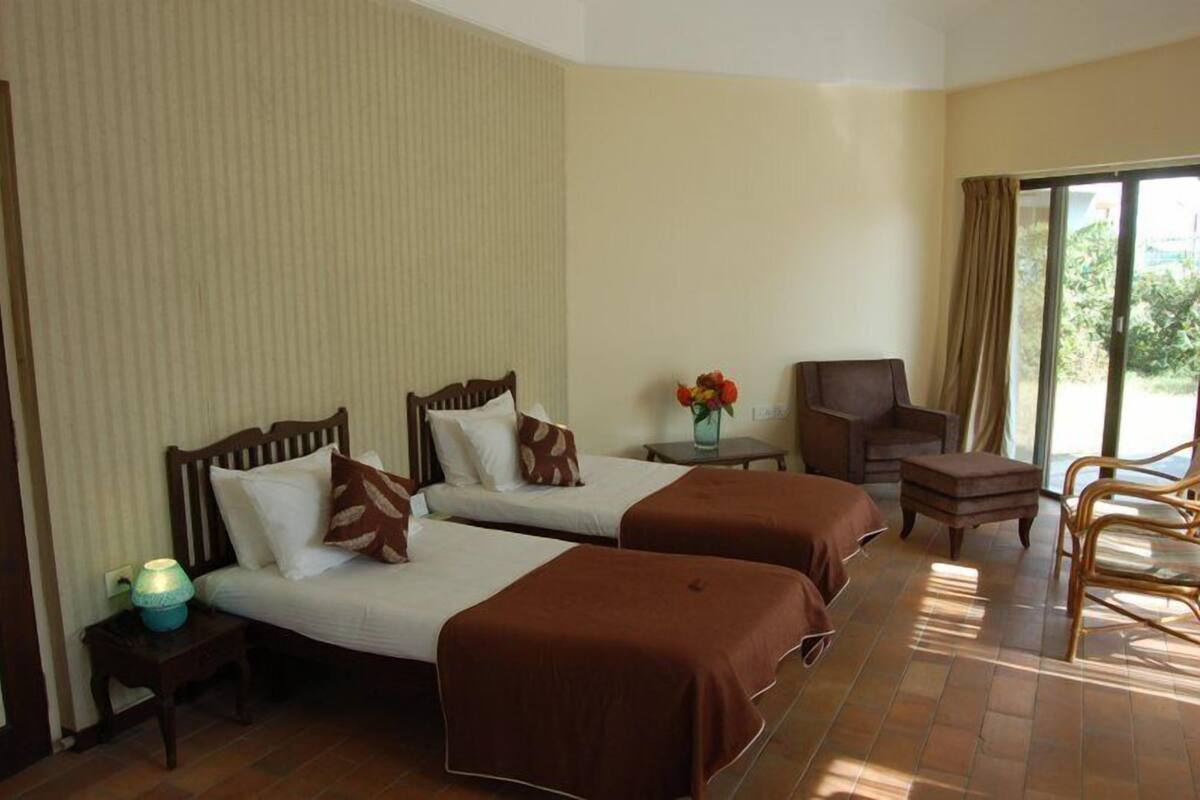 Une chambre d’hôtel avec deux lits, une petite table ornée d’un vase de fleurs, une chaise et une vue sur l’extérieur grâce à une grande fenêtre.