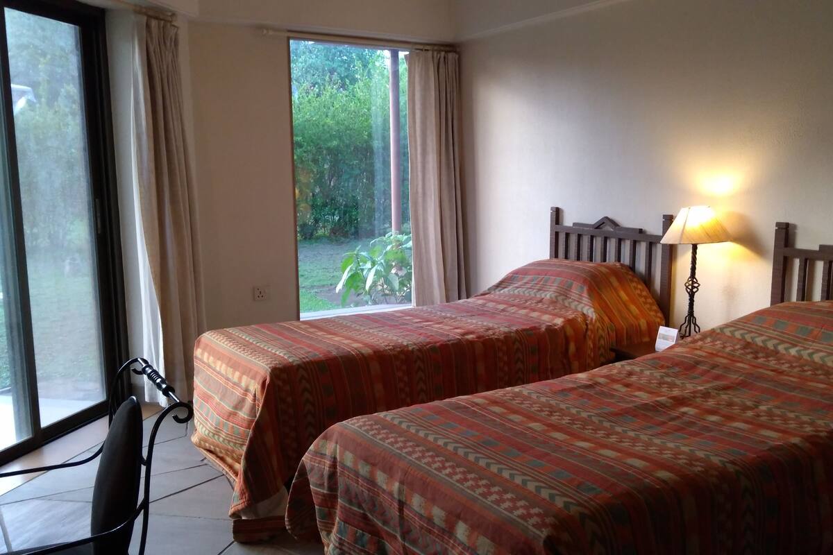 Une chambre avec deux lits, une fenêtre avec des rideaux, une lampe et une vue sur la verdure.