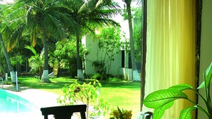 A dining area with a table set for two, chairs, and a view of a garden with palm trees and a pool. - The Meadows Resort and Spa (Chhatrapati Sambhajinagar)