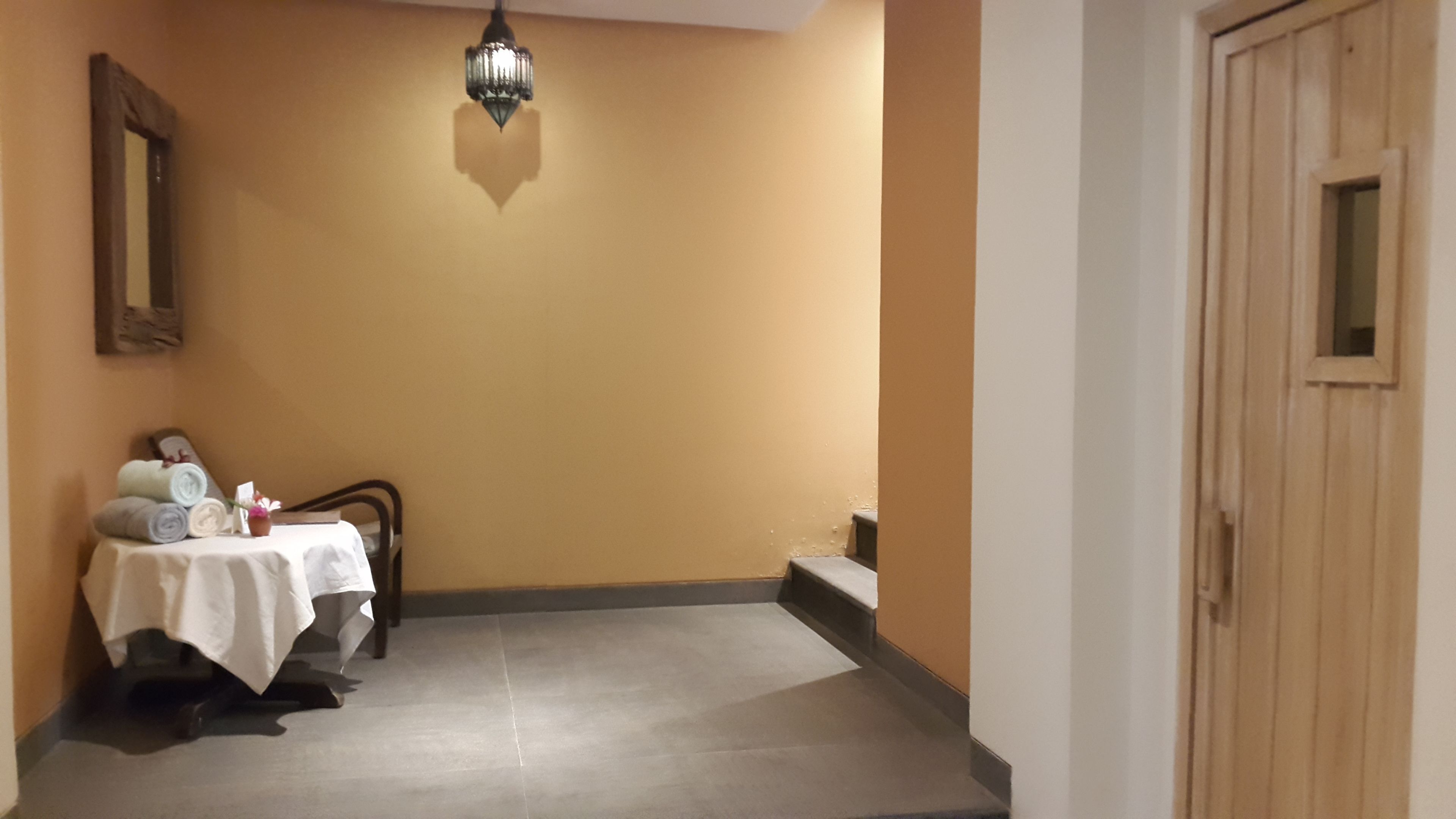 Habitación de hotel con cama, una mesa con toallas y un jarrón, una puerta de madera y una lámpara fijada a la pared.