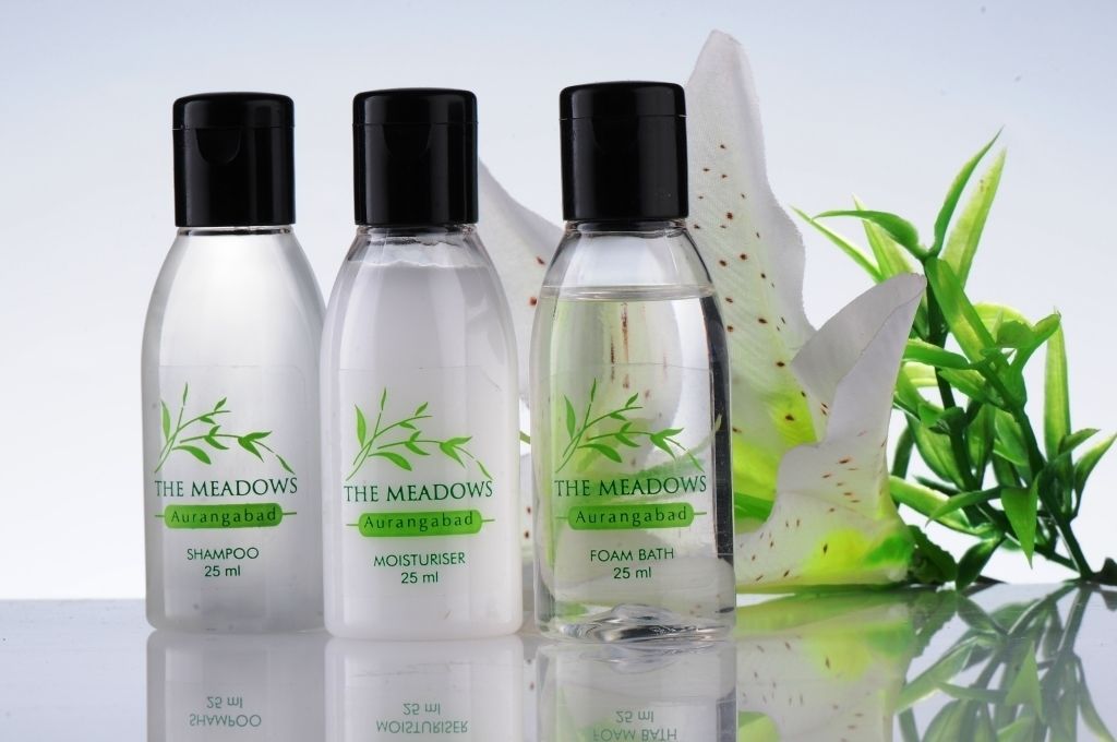 Tres botellas de champú, crema hidratante y espuma de baño The Meadows Aurangabad, cada una de 25 ml, con tapa negra, sobre un fondo blanco con una planta verde.