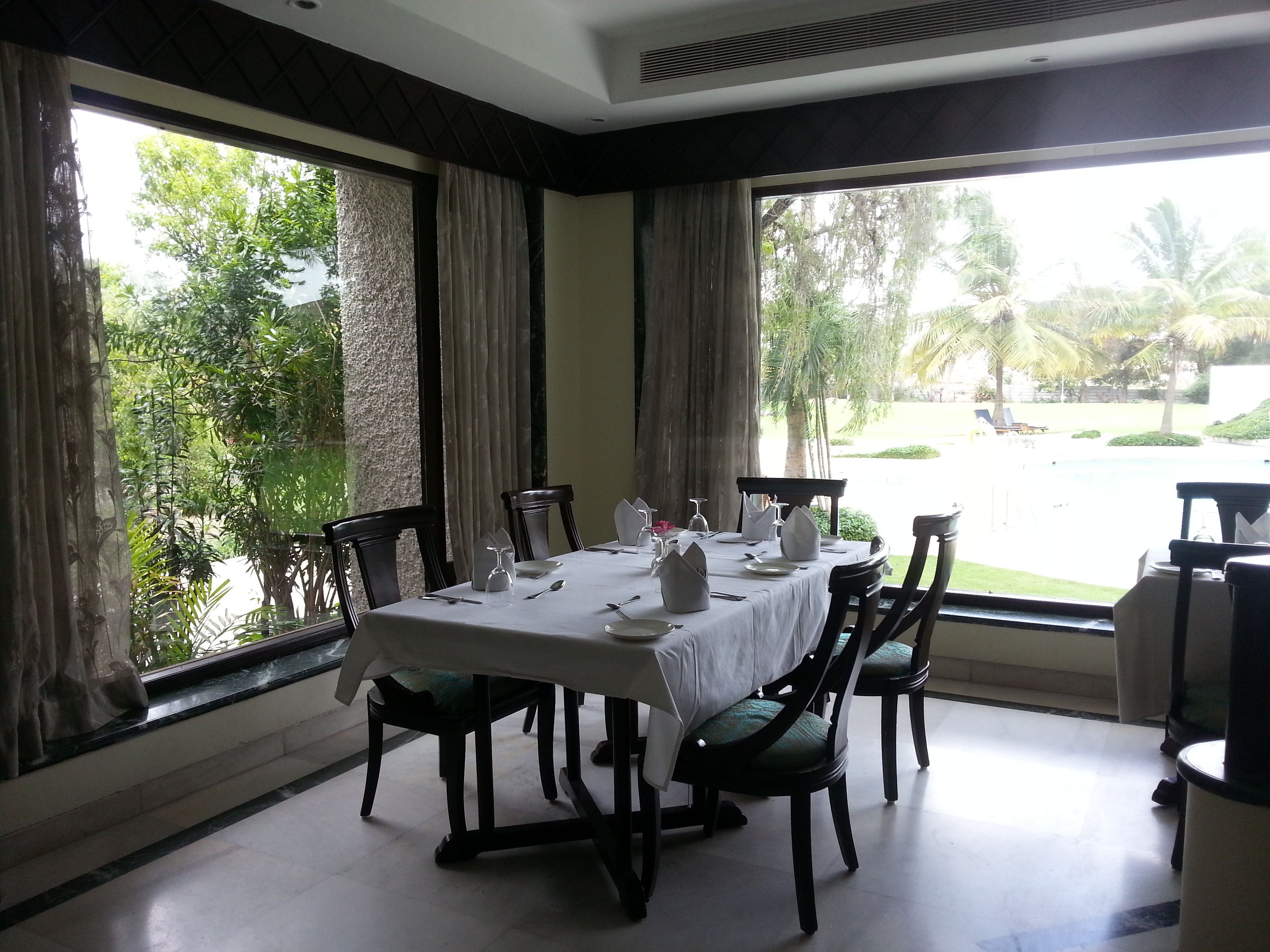 Un comedor con una mesa preparada para cuatro, ventanas grandes con cortinas y vistas a una piscina y follaje exterior.
