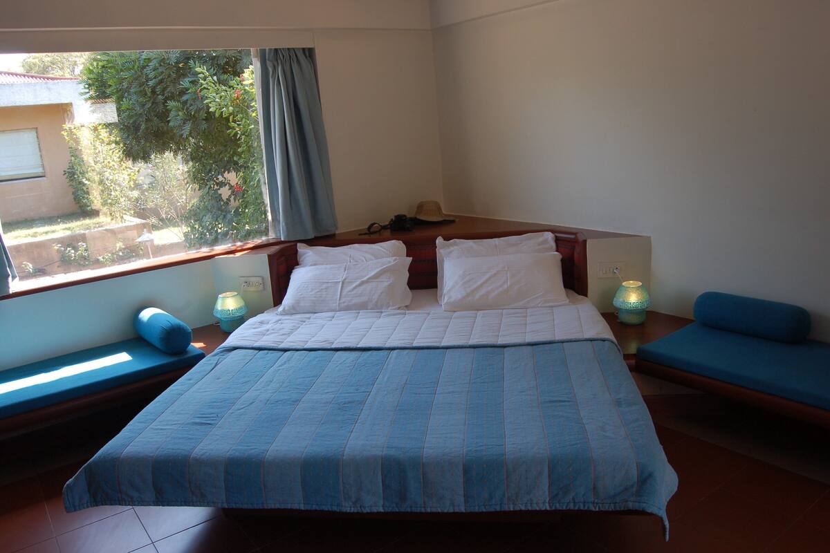 Une chambre avec un grand lit, deux tables de chevet, une fenêtre donnant sur un paysage verdoyant et un canapé bleu.