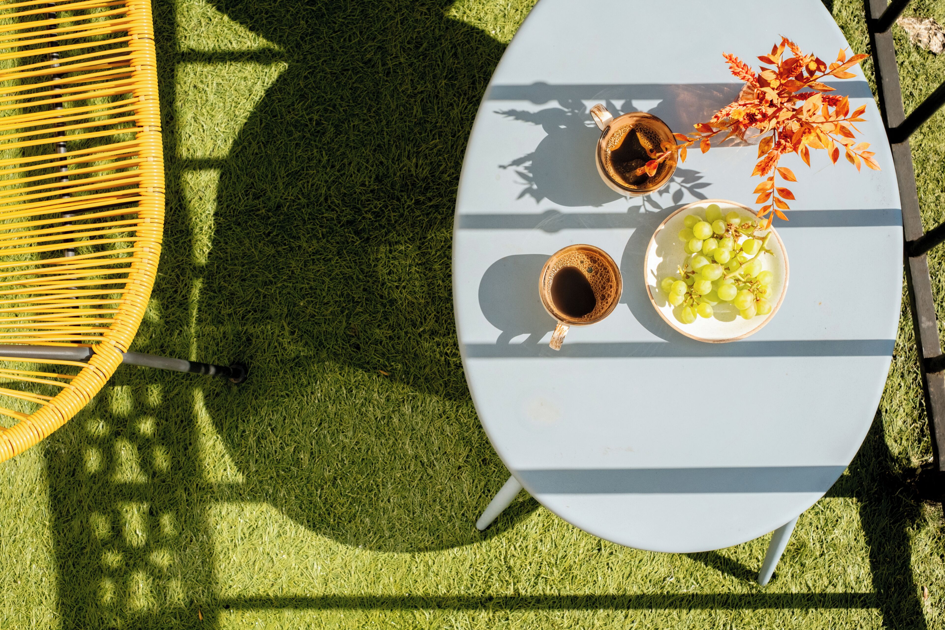 Une table ronde avec un plateau blanc, deux tasses de café, un bol de raisins verts et un vase rempli de feuilles orange.