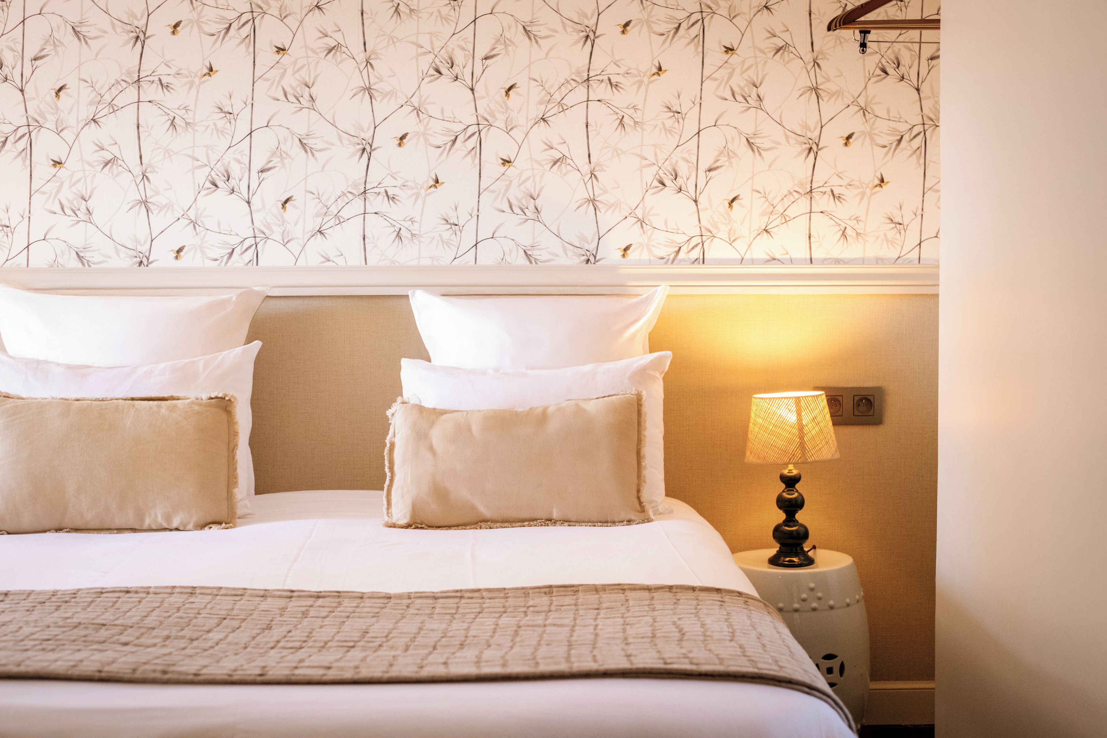 A neatly made bed with white and beige bedding, a wall with a bamboo leaf pattern, and a bedside lamp.