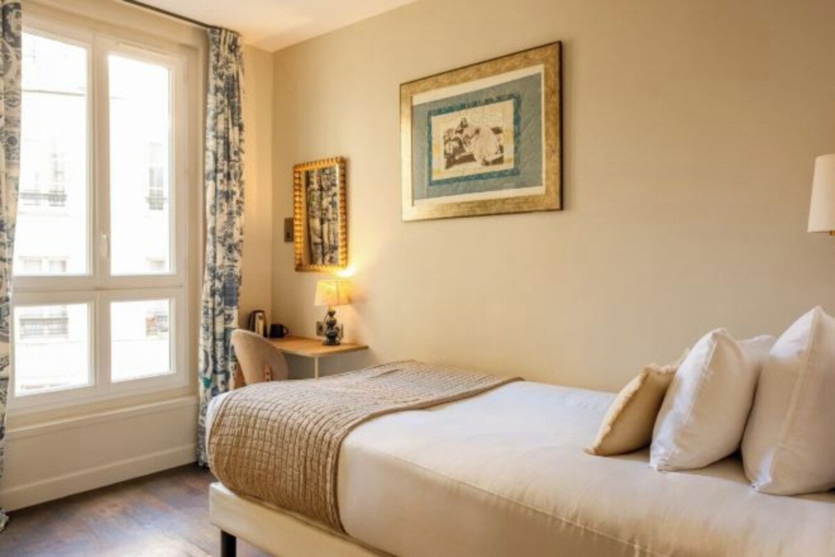 A hotel room with a bed, a desk, a lamp, a window with curtains, and a framed picture on the wall.