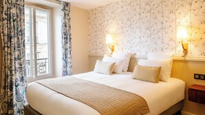A hotel room with a large bed, two bedside lamps, a window with curtains, and a floral wallpaper. - Hôtel R. Kipling by Happyculture (Paris)