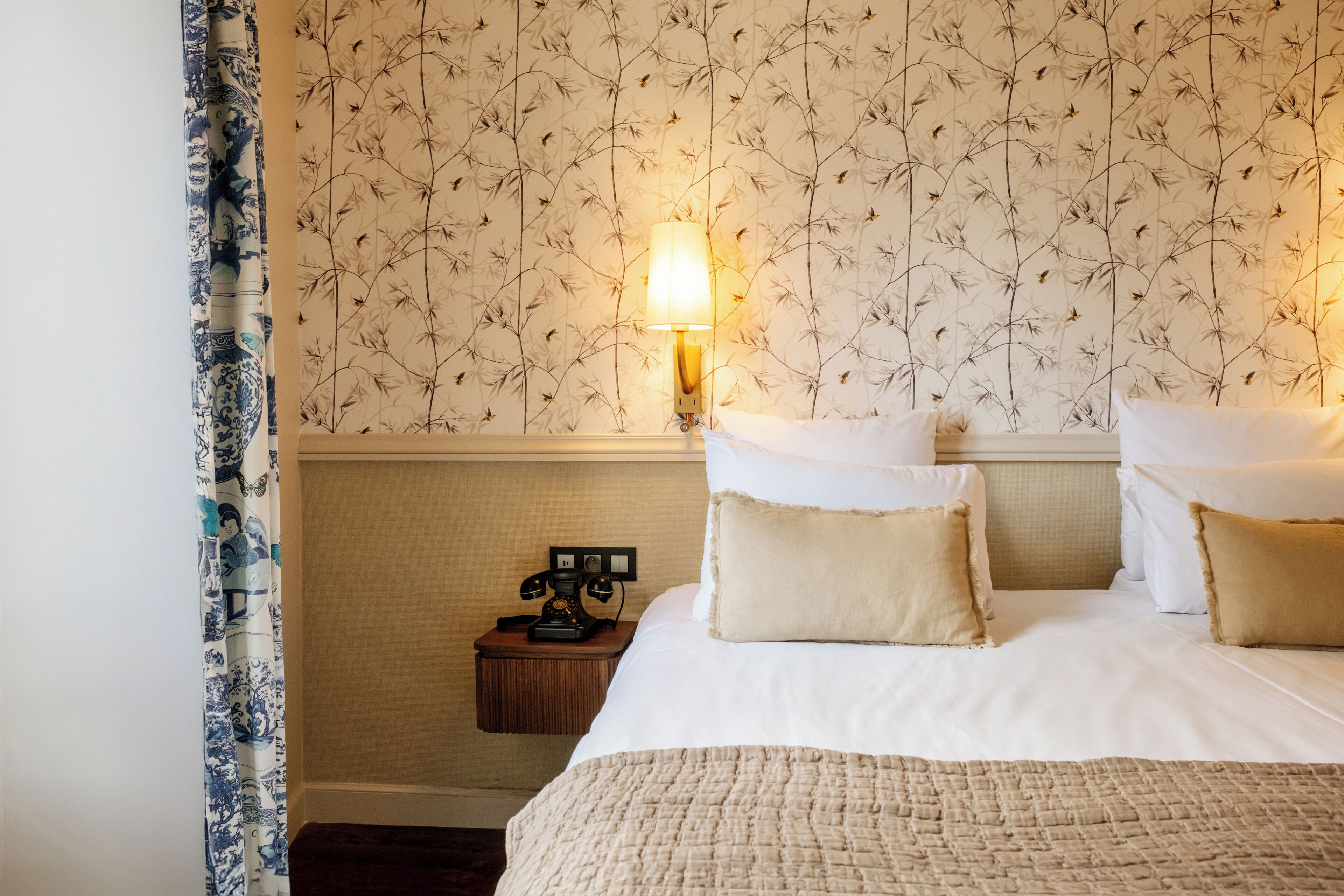 Habitación de hotel con cama, almohadas, mesita de noche y teléfono, y una pared con estampado floral.
