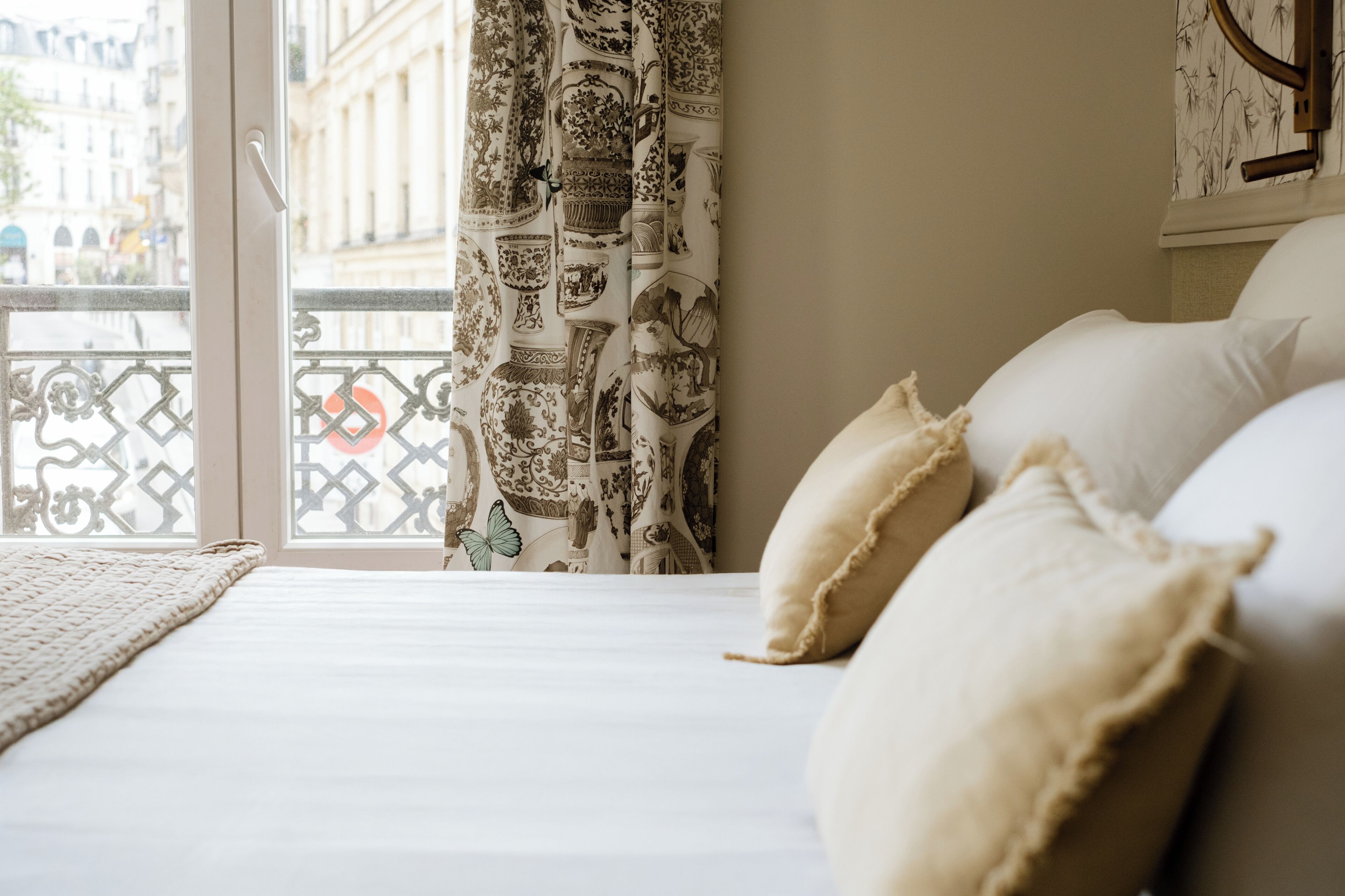 Un dormitorio con cama, almohadas, una ventana con cortinas estampadas y vista a edificios.