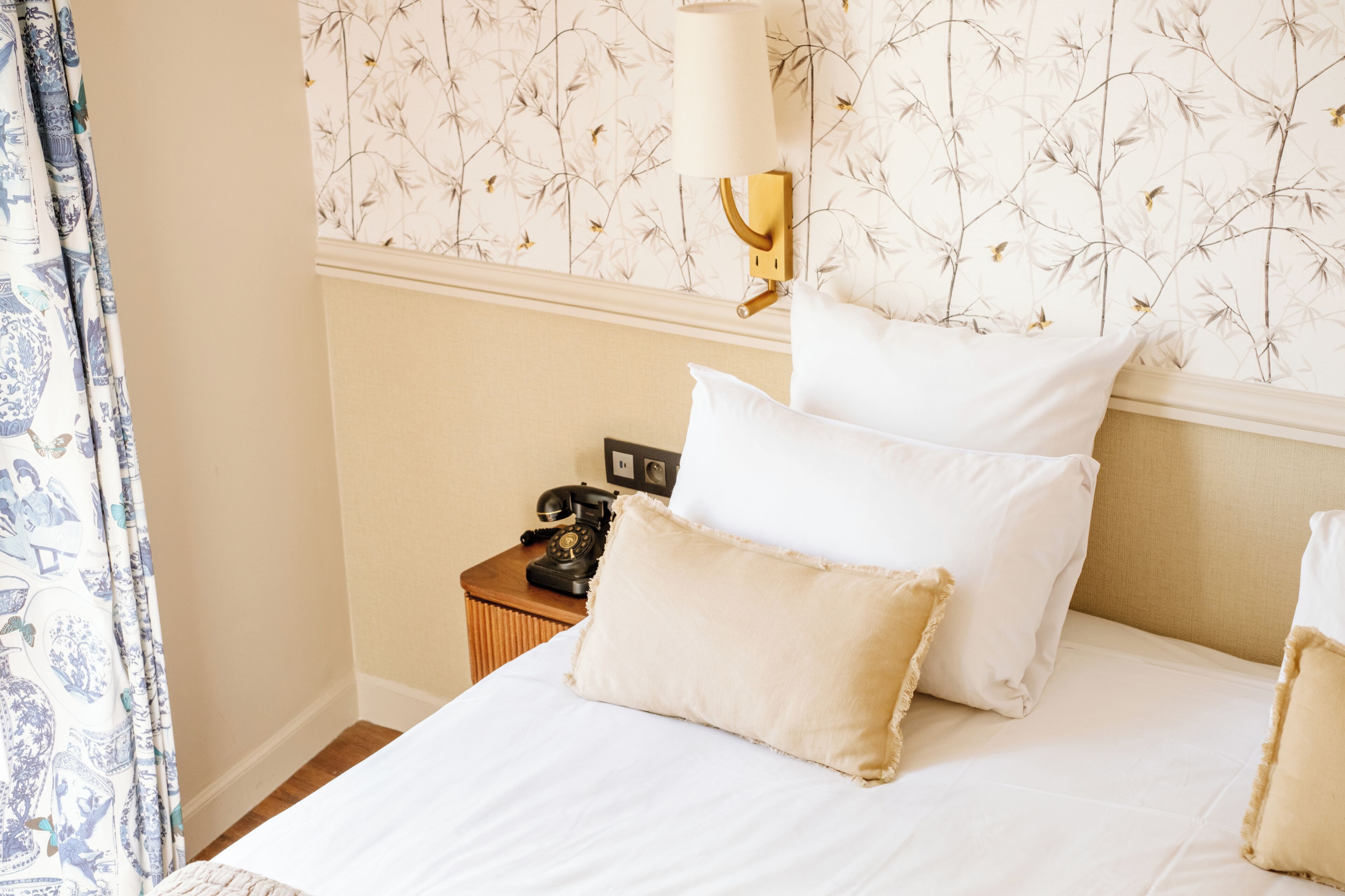 A hotel room with a bed, pillows, a nightstand with a telephone, and a wall with floral wallpaper.