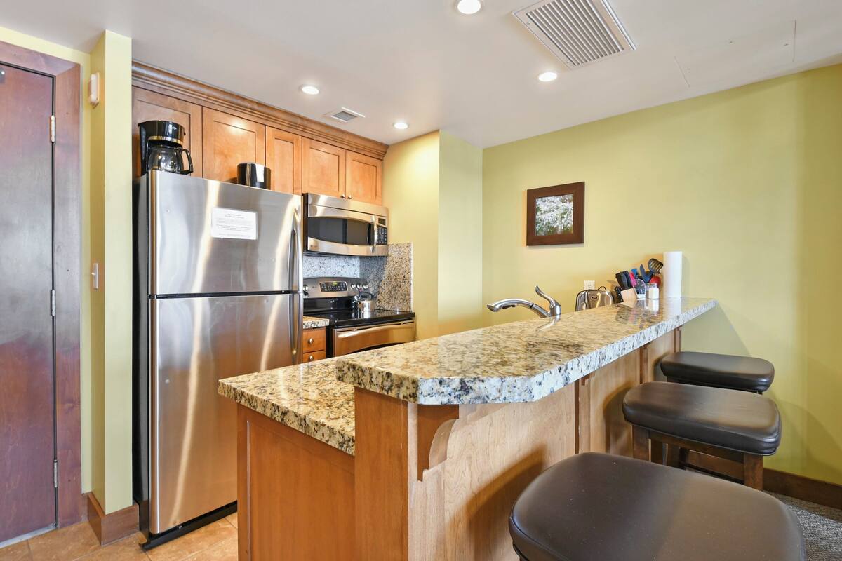 A kitchen with stainless steel appliances, wooden cabinets, granite countertops, and a bar area with stools.