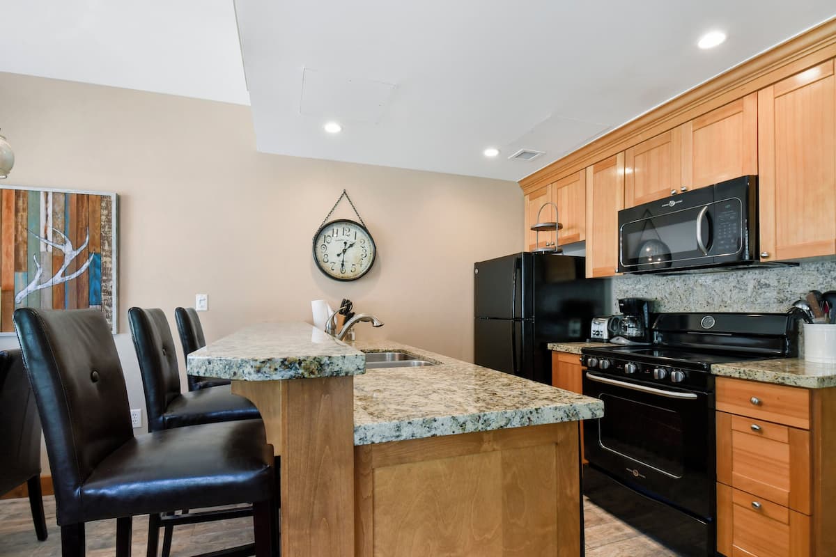 A kitchen with wooden cabinets, a granite countertop, a black refrigerator, a microwave, a stove, and a clock on the wall.