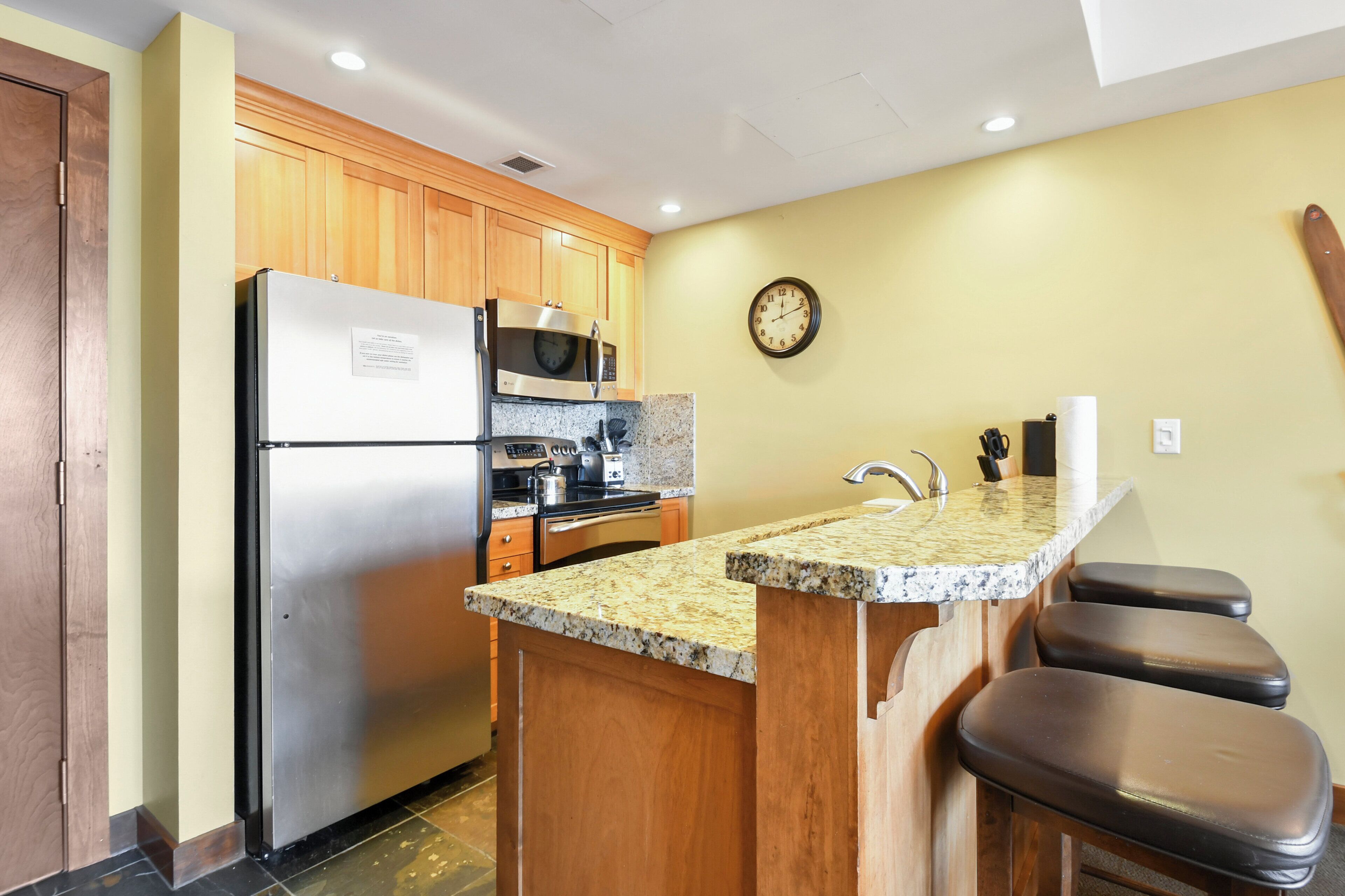 A kitchen with wooden cabinets, a stainless steel refrigerator, a microwave, an oven, a granite countertop, a sink, and a clock on the wall.
