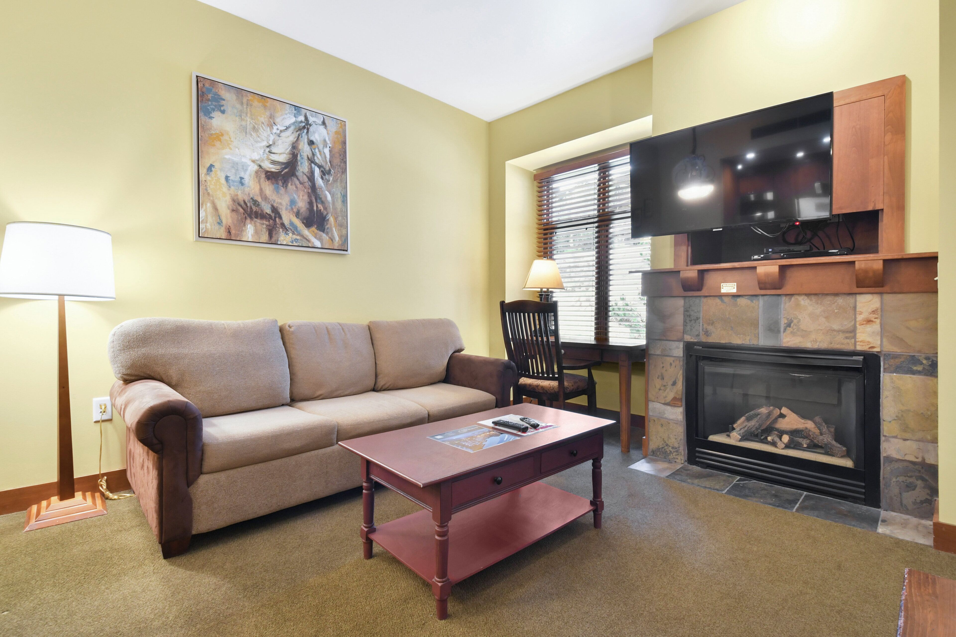 A living room with a beige sofa, a wooden coffee table, a fireplace, a flat-screen TV, and a painting on the wall.