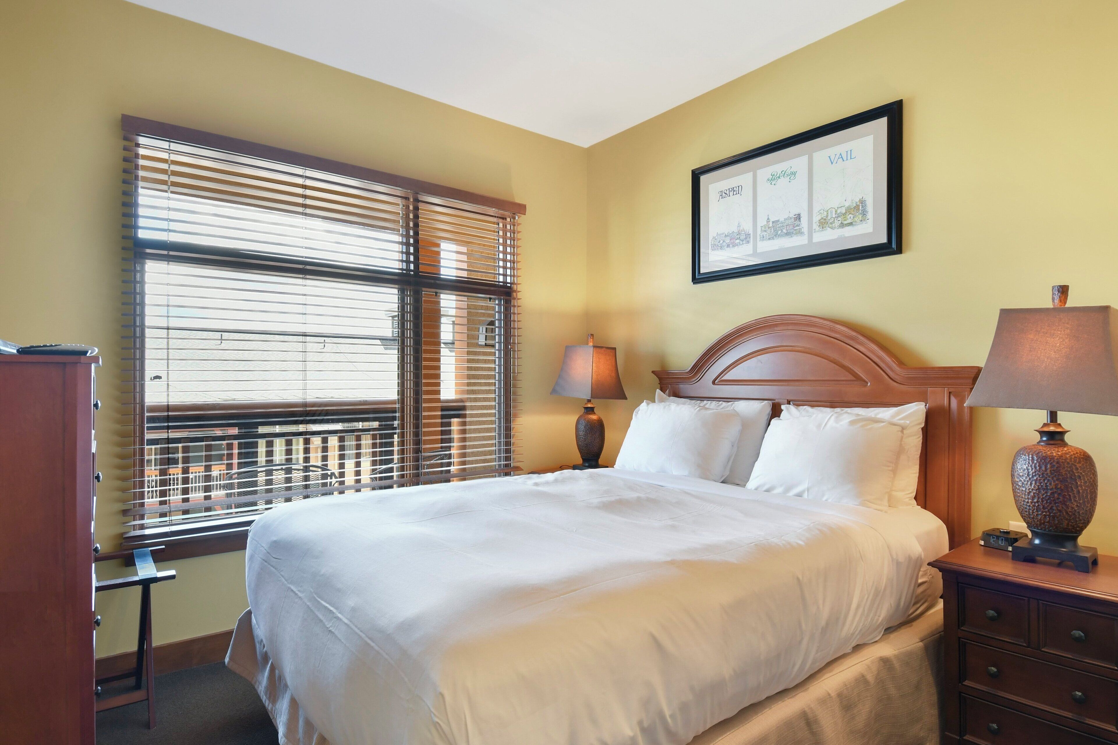 A hotel room with a bed, bedside table, lamp, and a window with blinds.