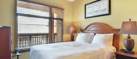 A hotel room with a bed, bedside table, lamp, and a window with blinds.