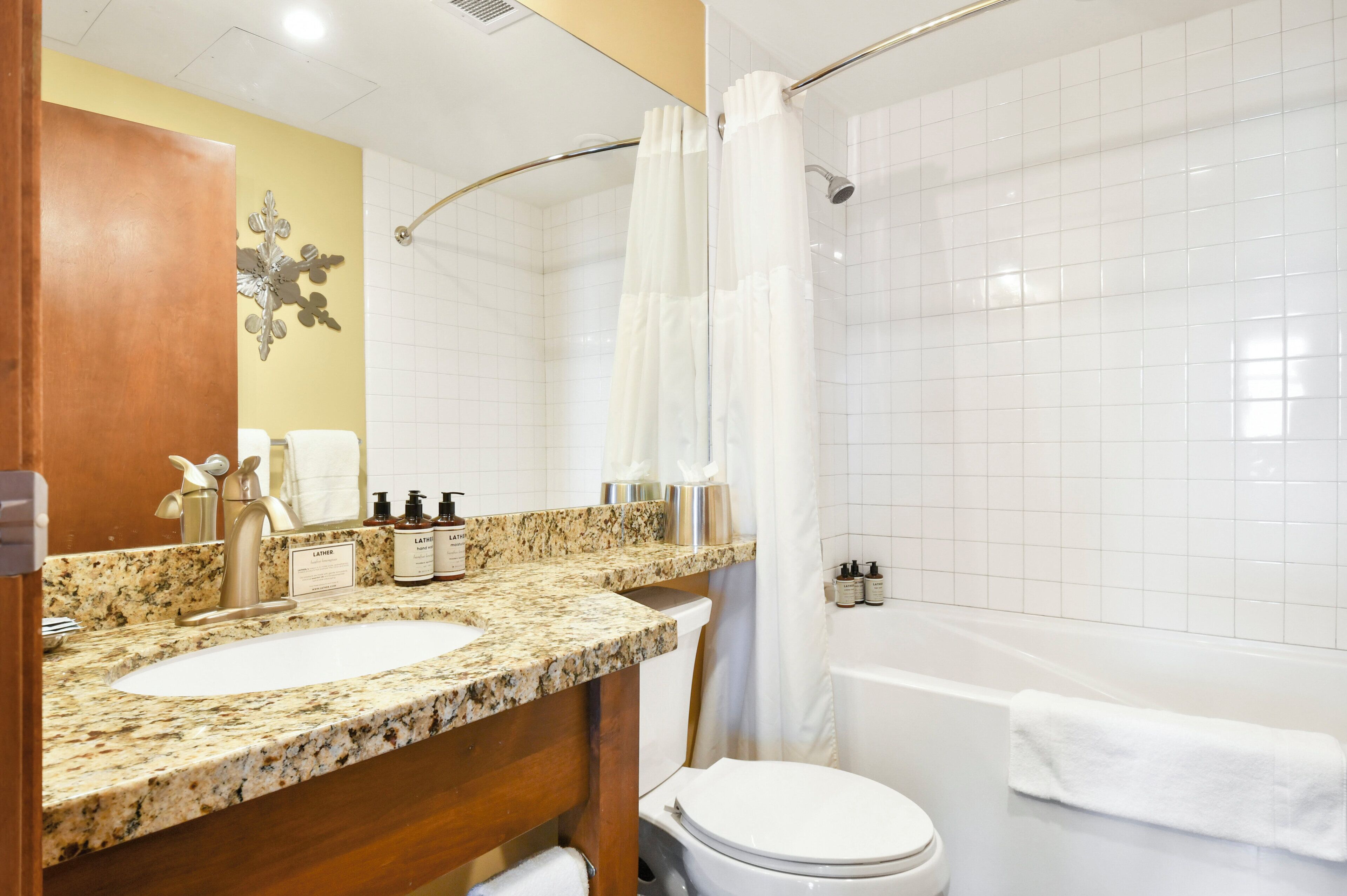 Een badkamer met een wastafel op een granieten blad, toilet, douche met een witte gordijn en een spiegel met een decoratief element aan de muur.