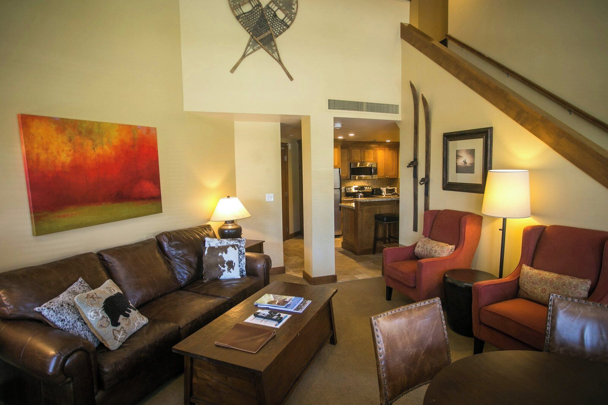 A living room with a brown leather sofa, a coffee table, and two armchairs. There is a painting on the wall and a kitchen visible through an open door.