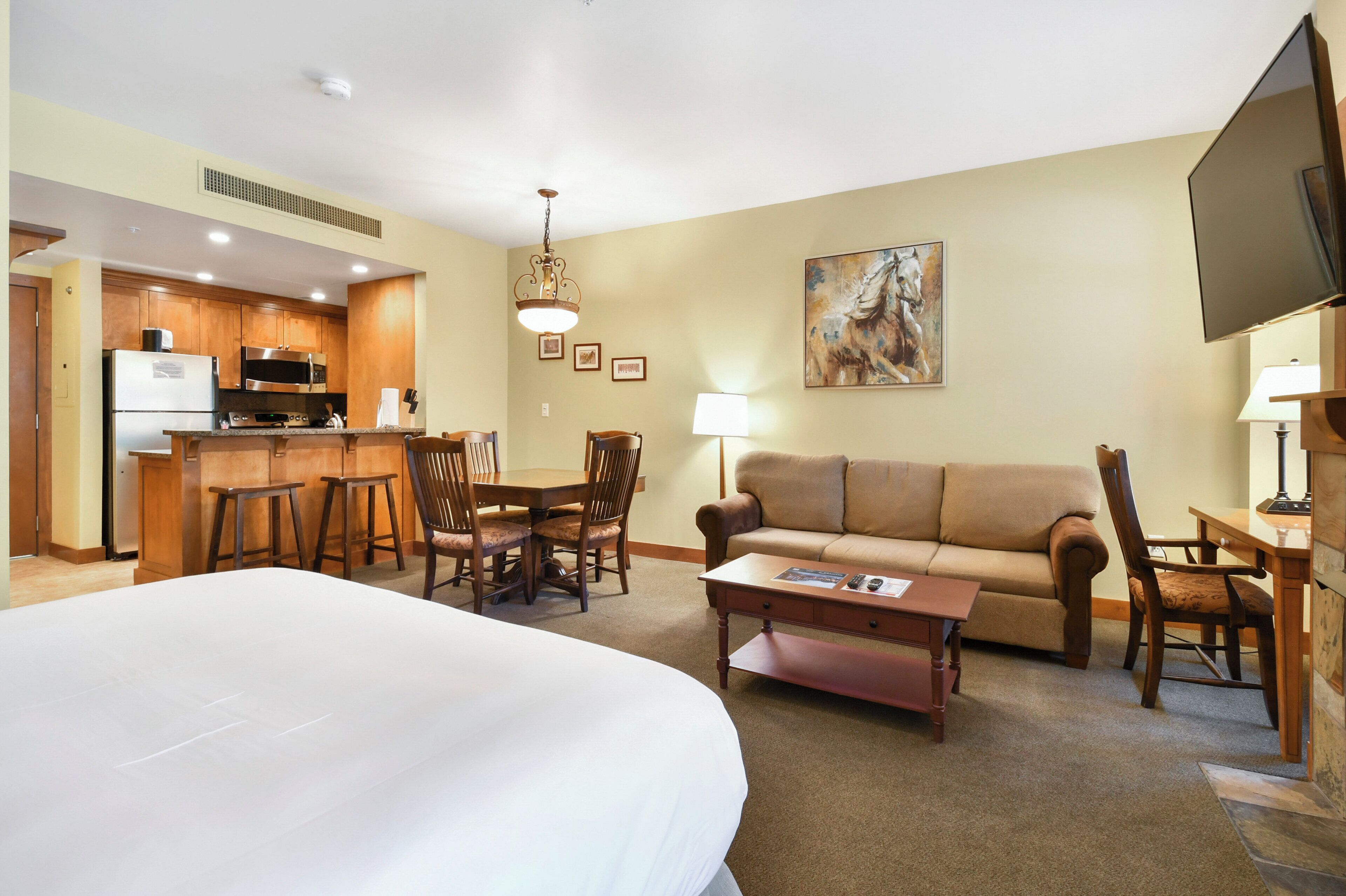 A hotel room with a large bed, a sofa, a coffee table, a dining area with chairs, a kitchen with appliances, and a TV.