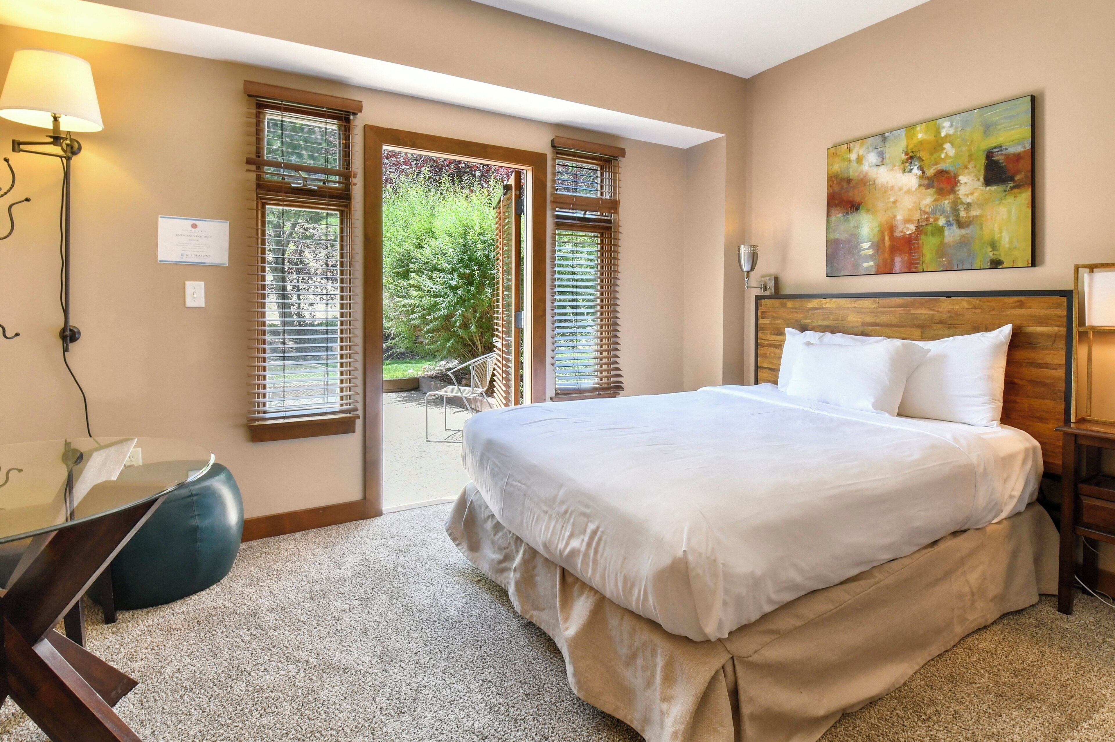 Une chambre avec un grand lit, un bureau et une chaise. Il y a deux fenêtres avec des stores et un tableau au mur.