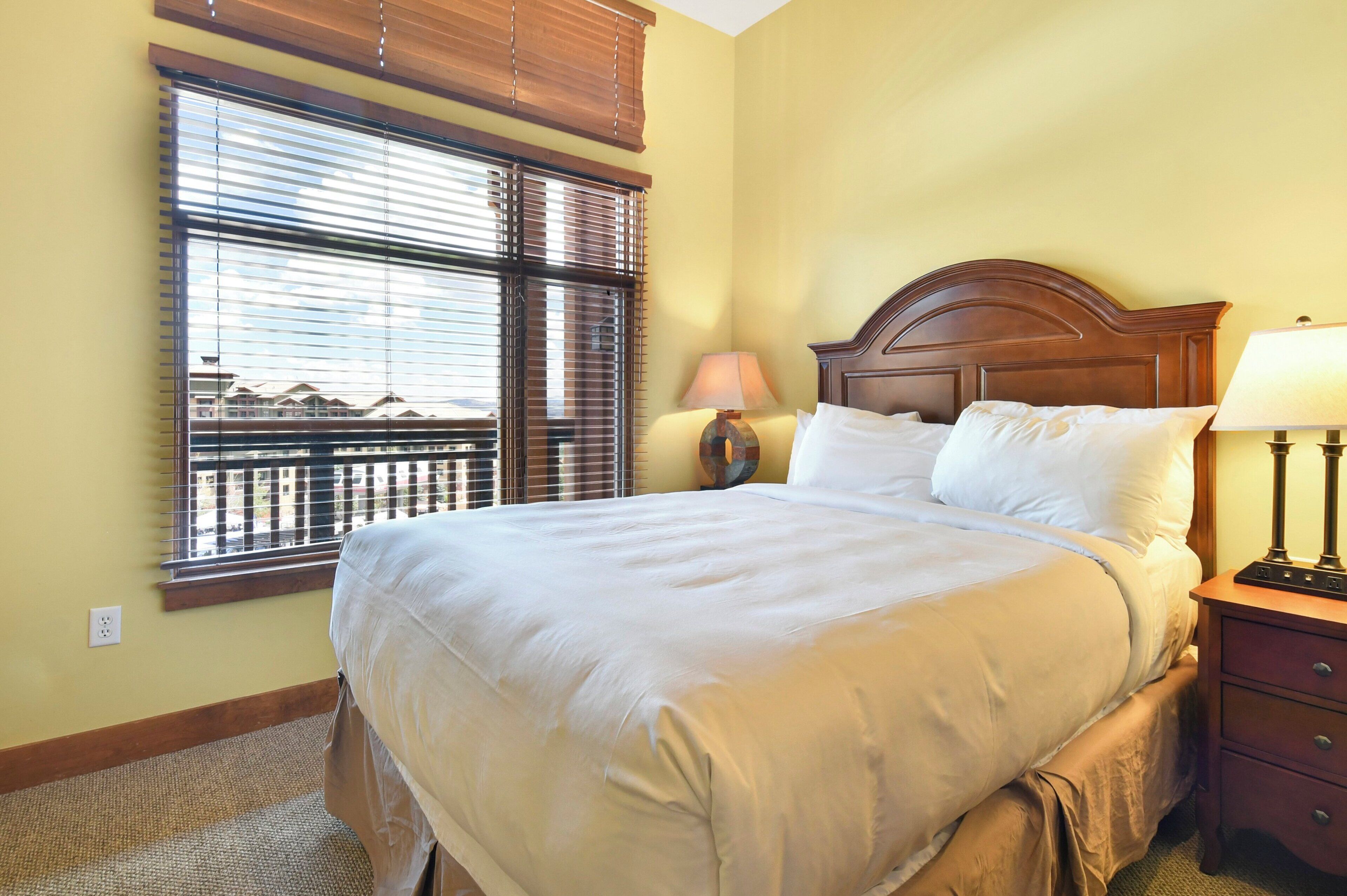 A neatly made bed with a wooden headboard, a bedside lamp, and a window with blinds.