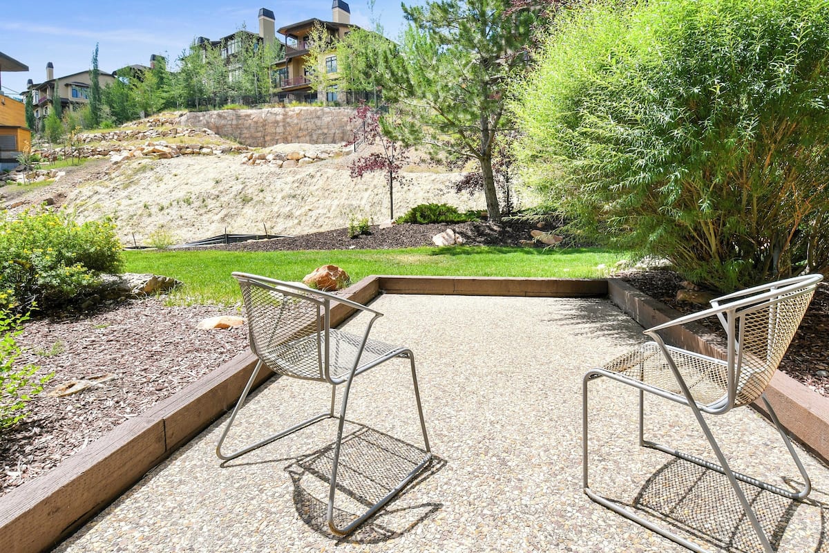 A patio area with two metal chairs, a stone retaining wall, and a grassy slope with trees and a house in the background.