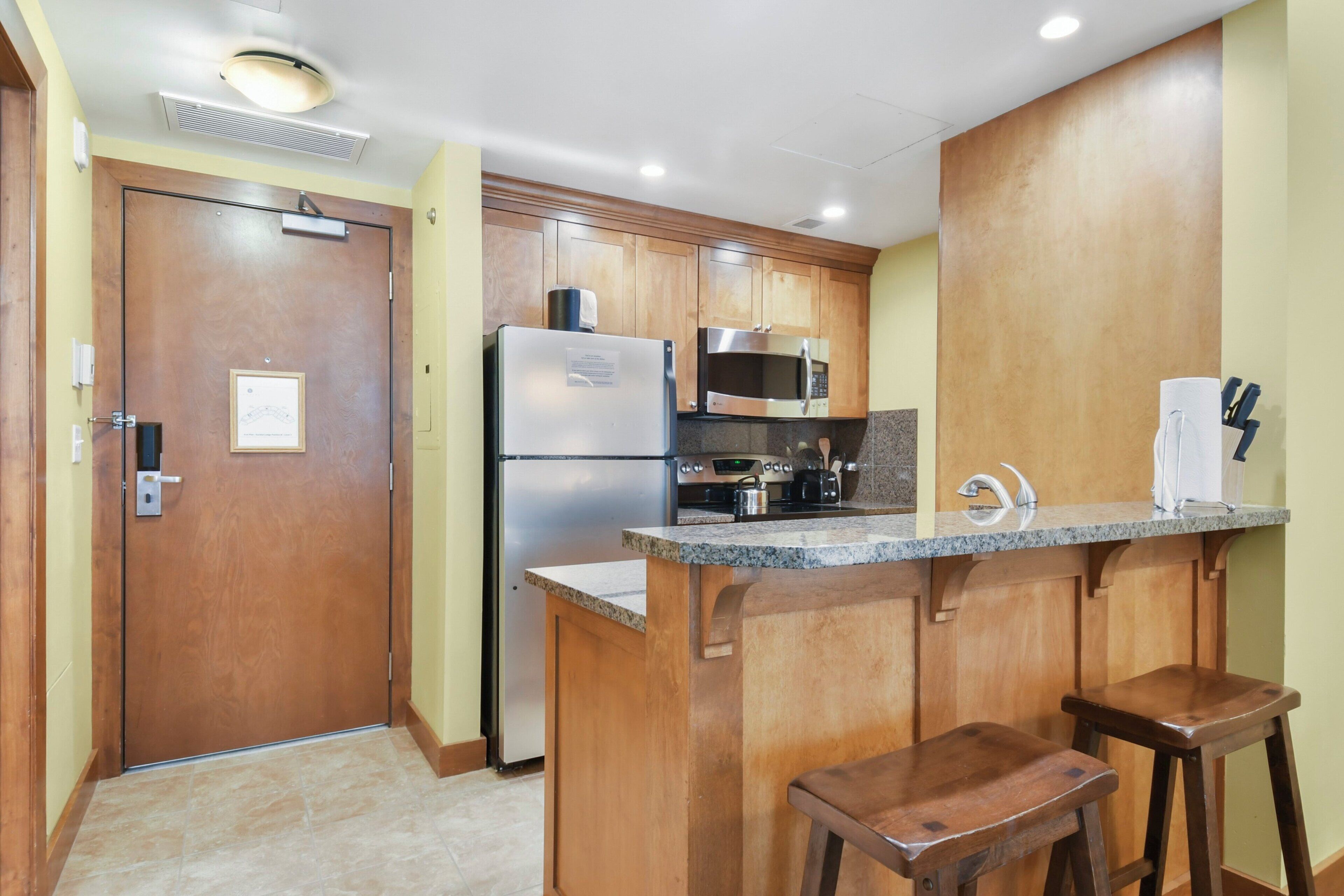 A compact kitchen with wooden cabinets, a stainless steel refrigerator, a microwave, and a granite countertop. There are two wooden stools at the counter.