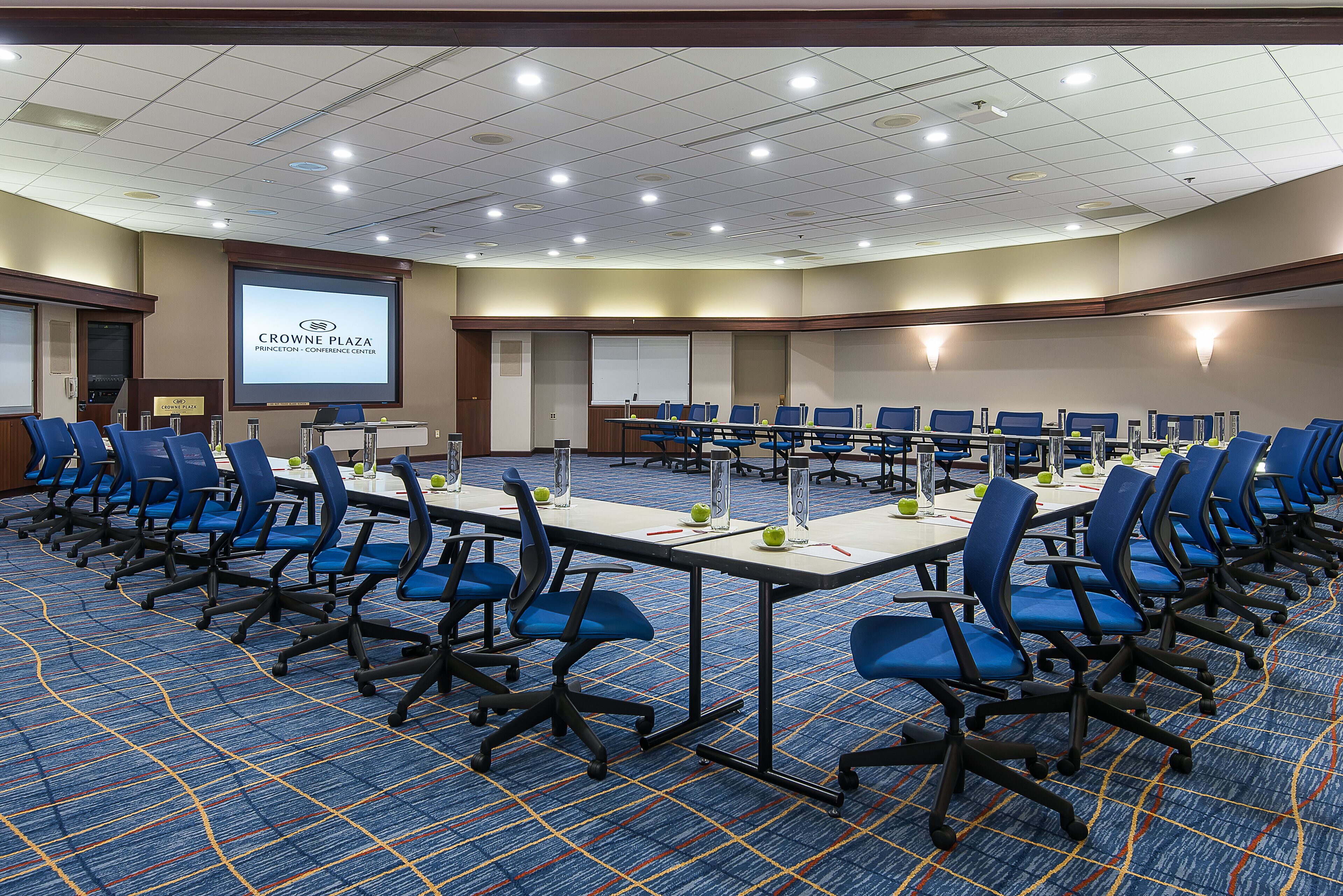 A conference room with rows of blue chairs, a large screen displaying 'Crowne Plaza', and a carpeted floor.