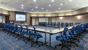 A conference room with rows of blue chairs, a large screen displaying 'Crowne Plaza', and a carpeted floor.