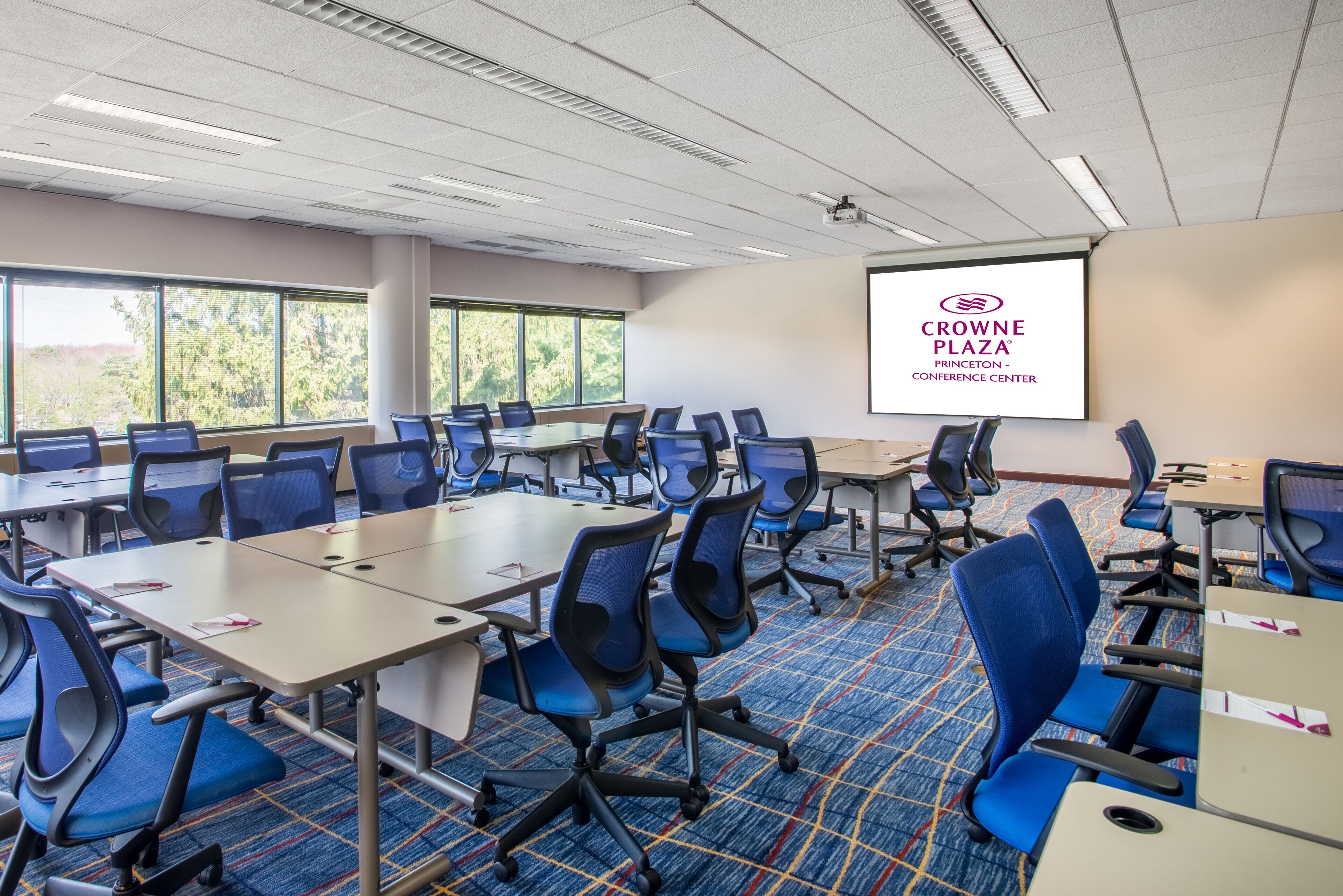 A conference room with multiple tables and chairs, large windows, and a projector screen displaying the Crowne Plaza logo.