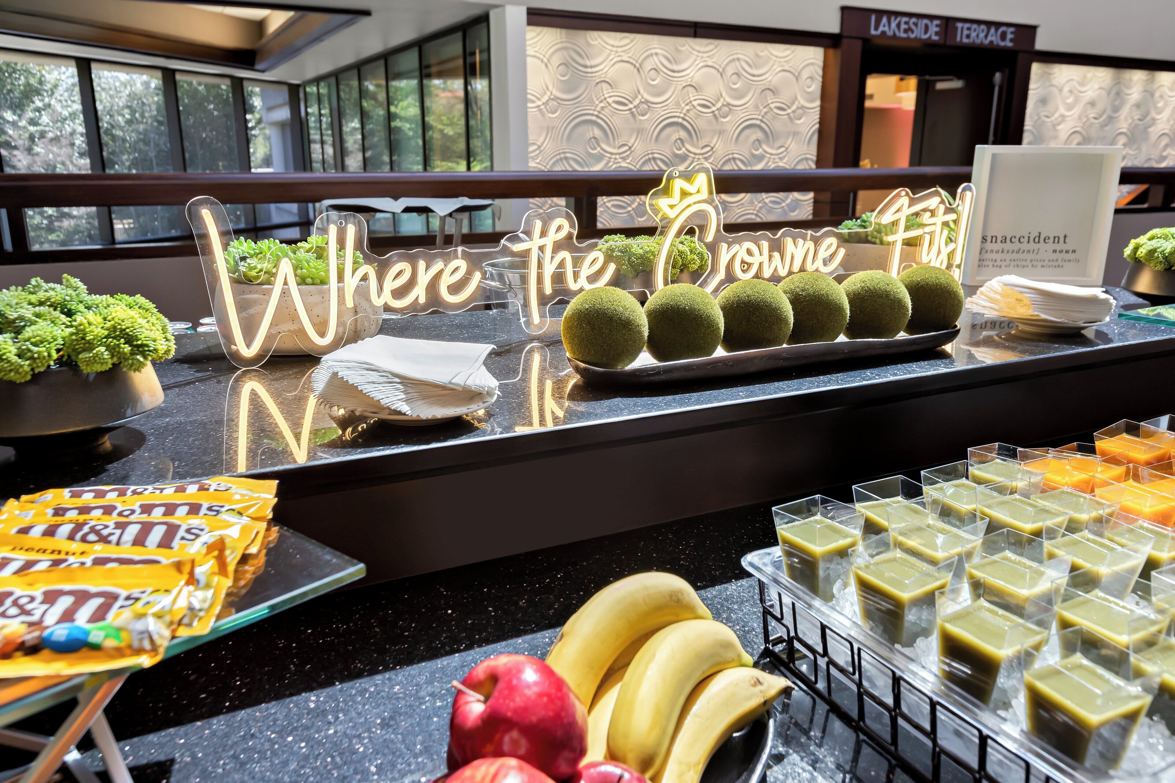 Una mesa de buffet con un letrero de neón que dice "Where the Crown Fits!" y diferentes alimentos como plátanos, manzanas y aperitivos envasados.