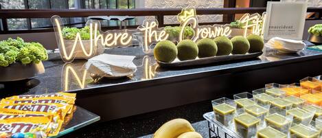 Una mesa de buffet con un letrero de neón que dice "Where the Crown Fits!" y diferentes alimentos como plátanos, manzanas y aperitivos envasados.