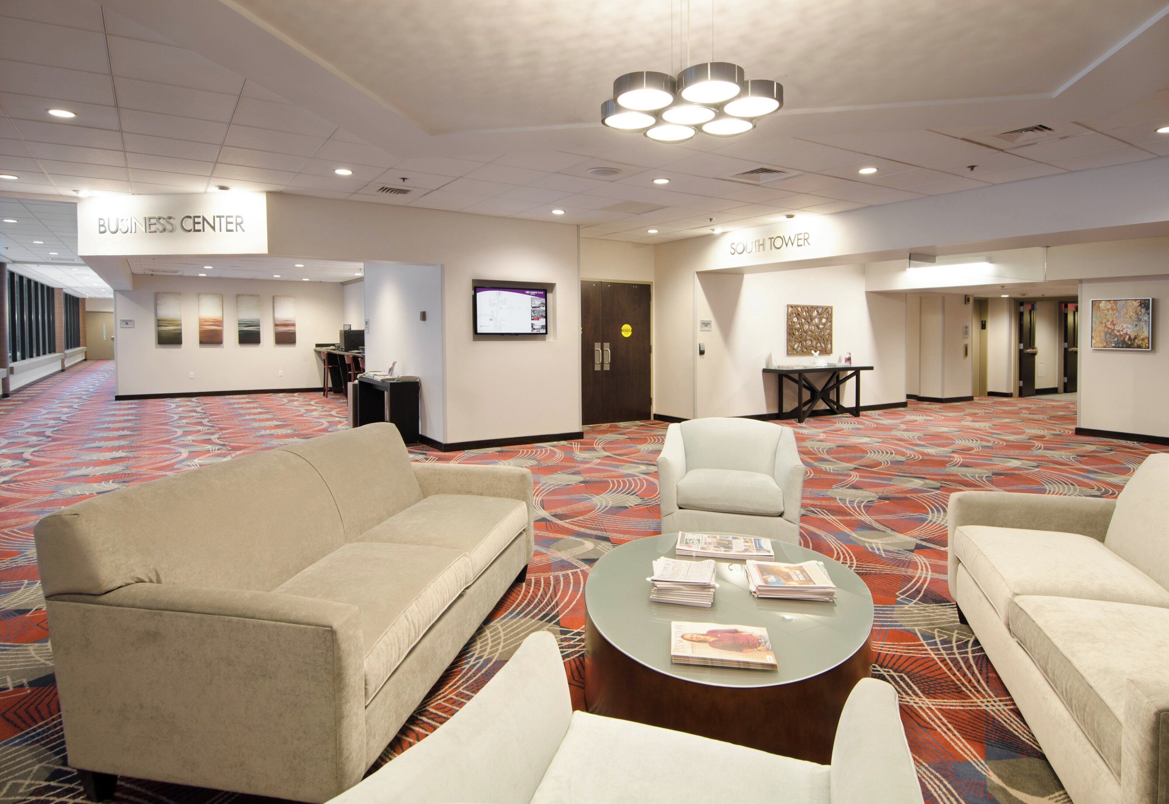 A spacious business center with a large couch, a round table with magazines, and a television on the wall.