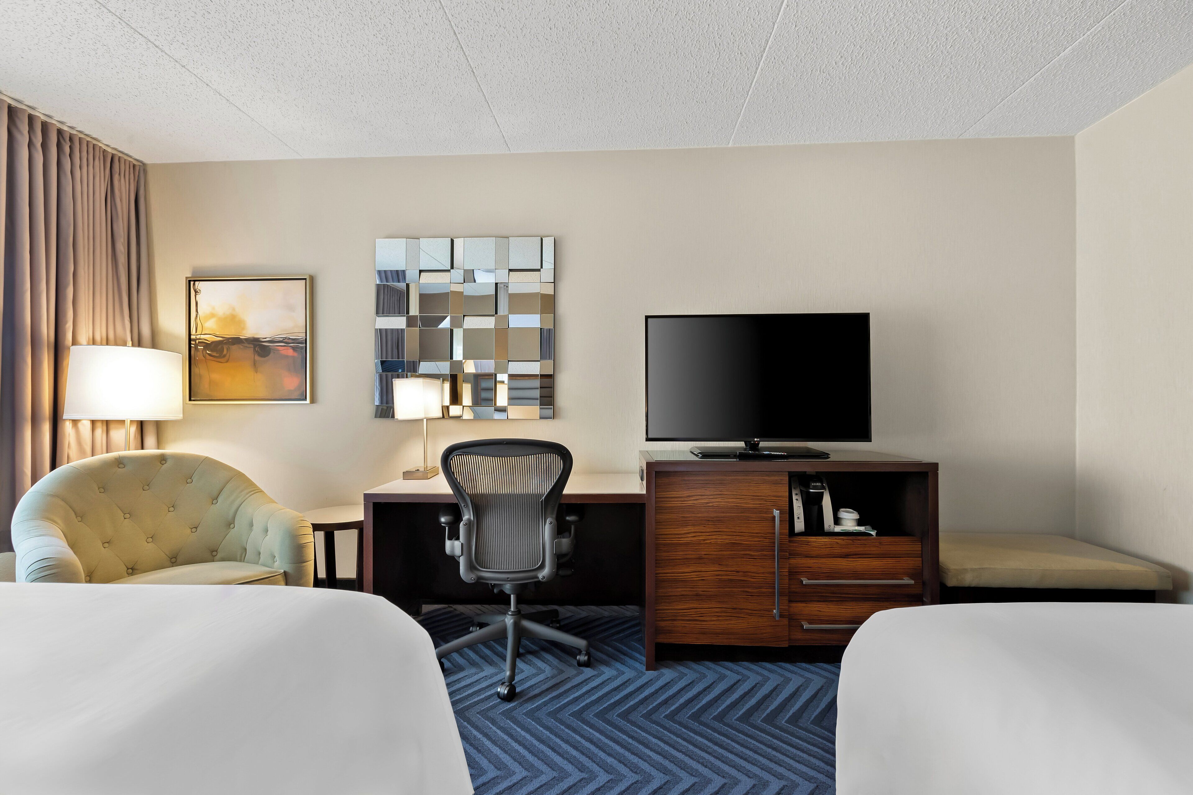 A hotel room with two beds, a desk with a chair, a television, a lamp, and a decorative mirror.