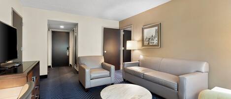 A hotel room with a TV, a sofa, a chair, a coffee table, and a lamp.