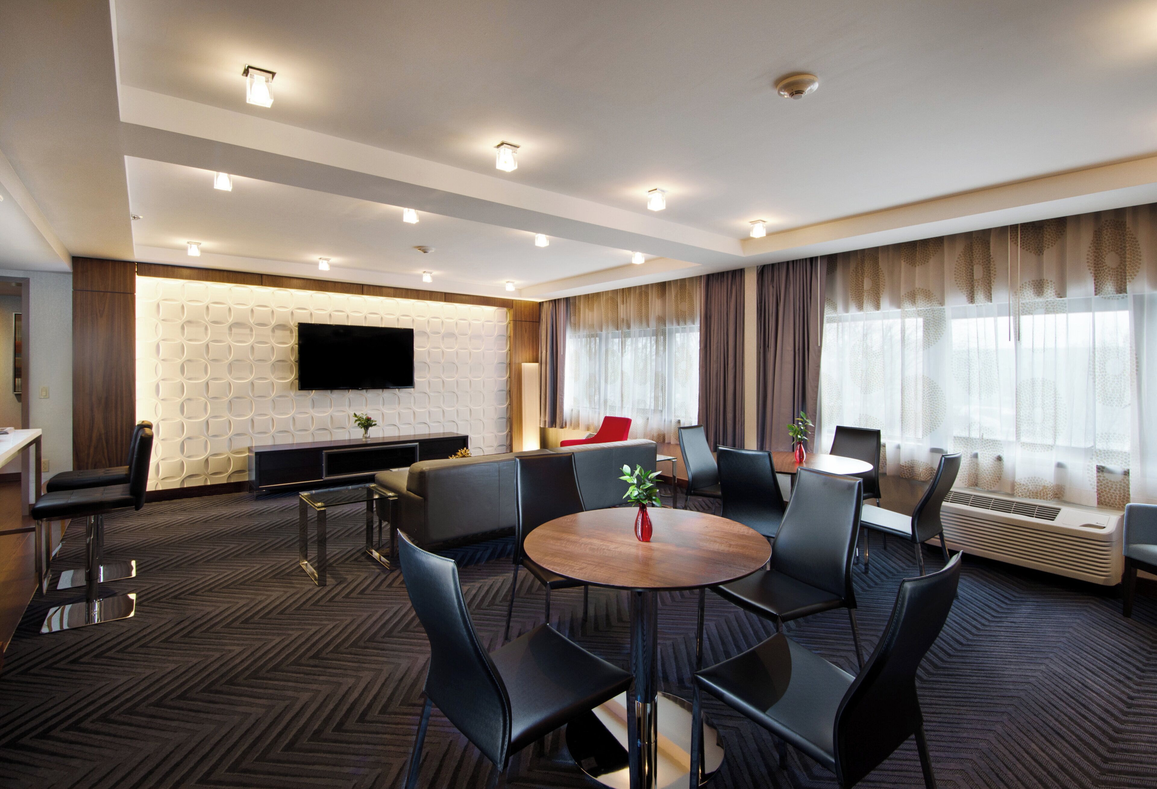 Habitación de hotel con televisión de pantalla plana montada en una pared texturizada, un sofá, una mesa de comedor con sillas y un ventanal con cortinas.
