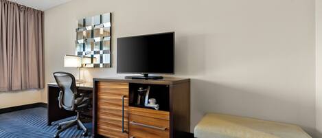 Quarto de hotel com escrivaninha, cadeira, televisão e um banco.