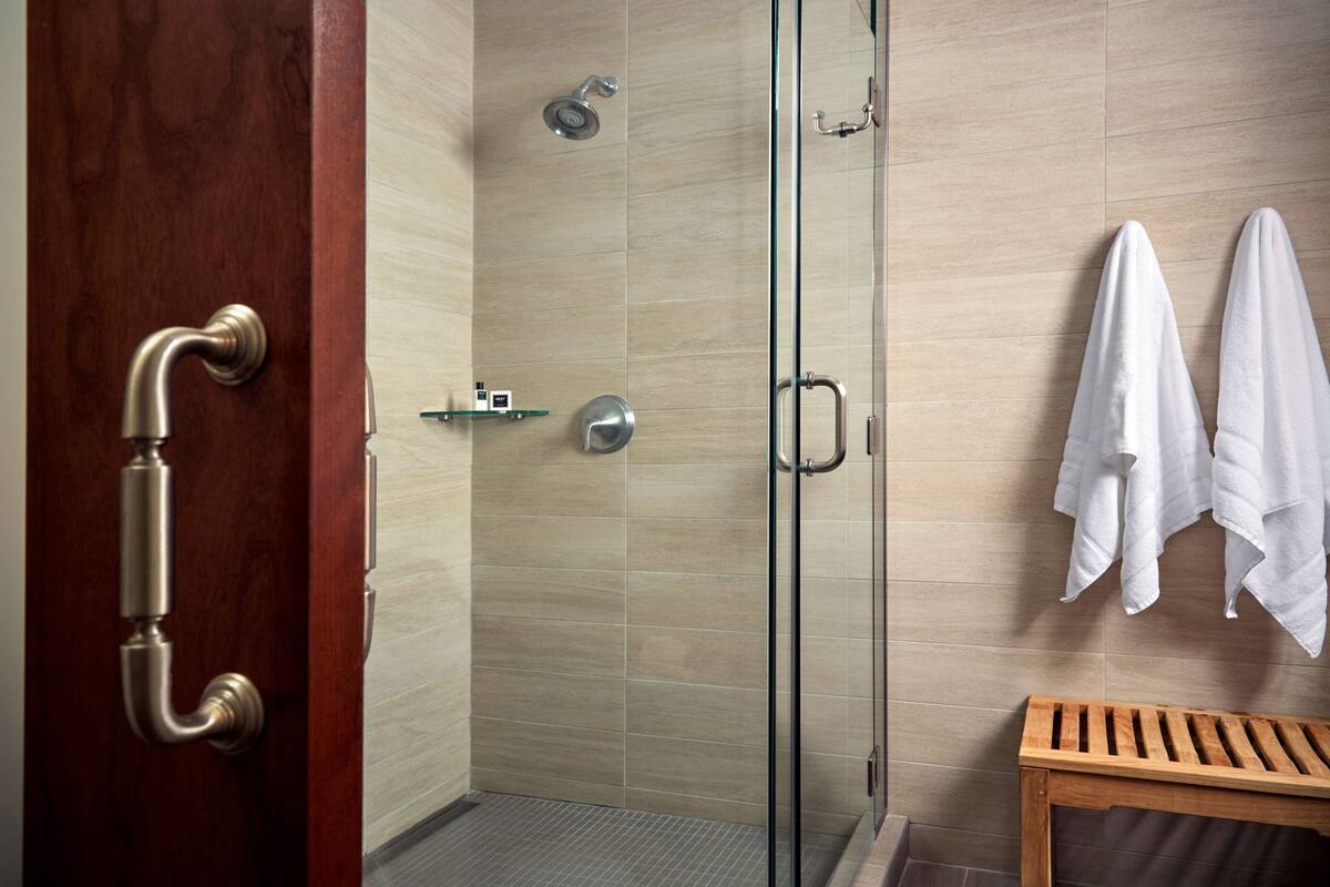 A modern bathroom with a glass shower enclosure, a wooden door with a silver handle, and a wooden bench.