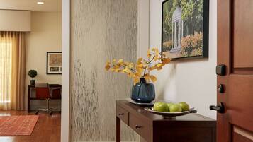 A modern interior with a wooden floor, a desk with a vase and apples, a door with a keypad, and a framed picture on the wall.
