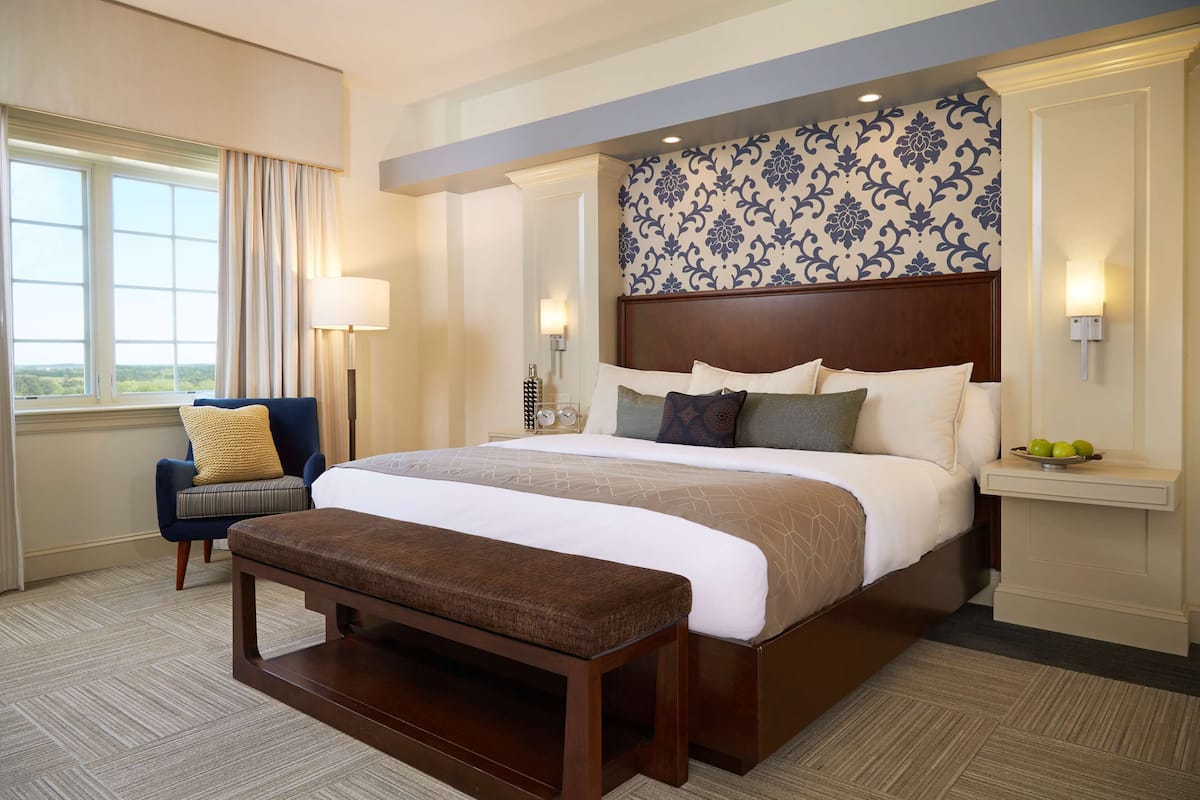 A hotel room with a large bed, a bench, a chair, a nightstand, and a view of the outdoors.