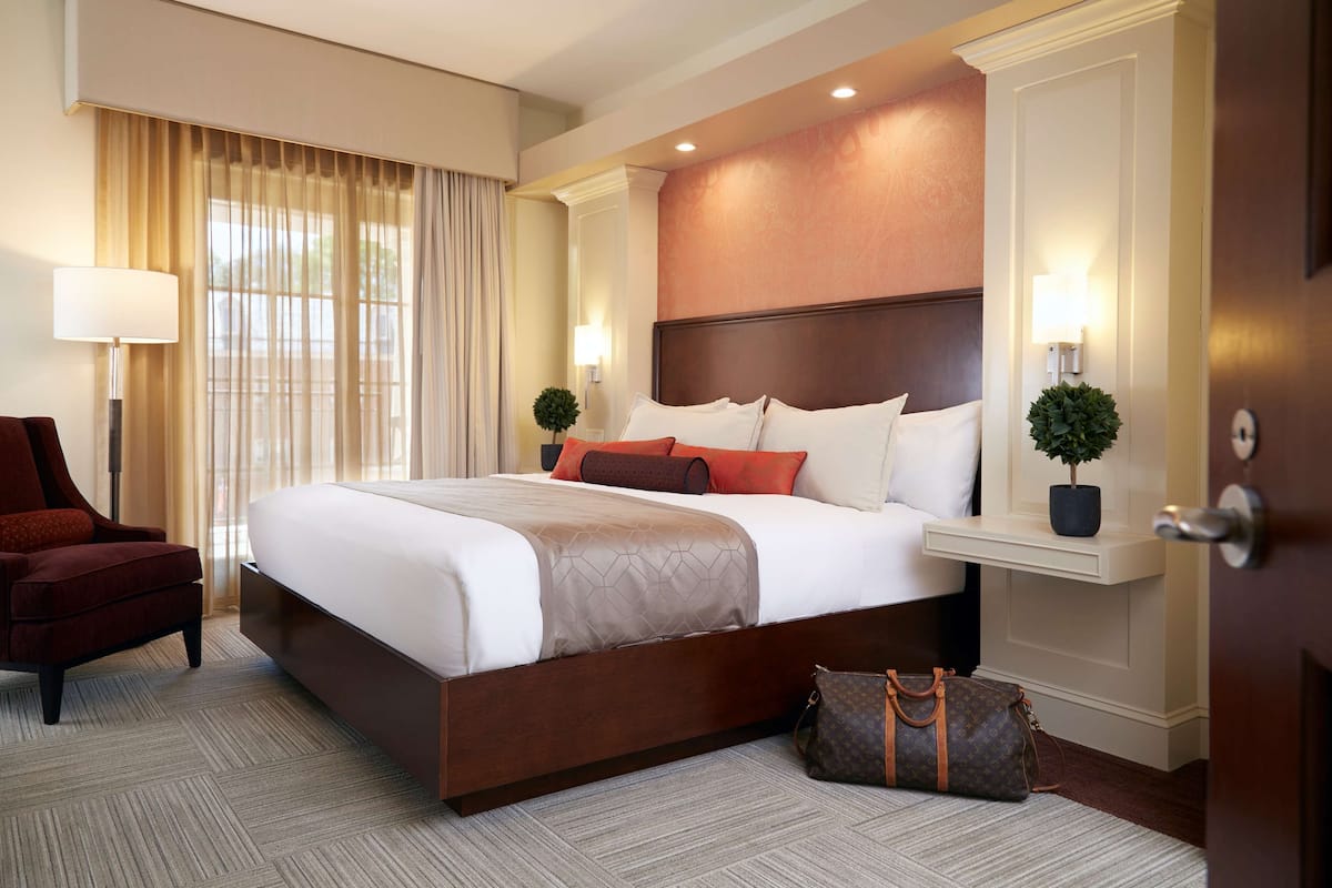 A hotel room with a large bed, a red armchair, a nightstand with a lamp, and a door with a handle.